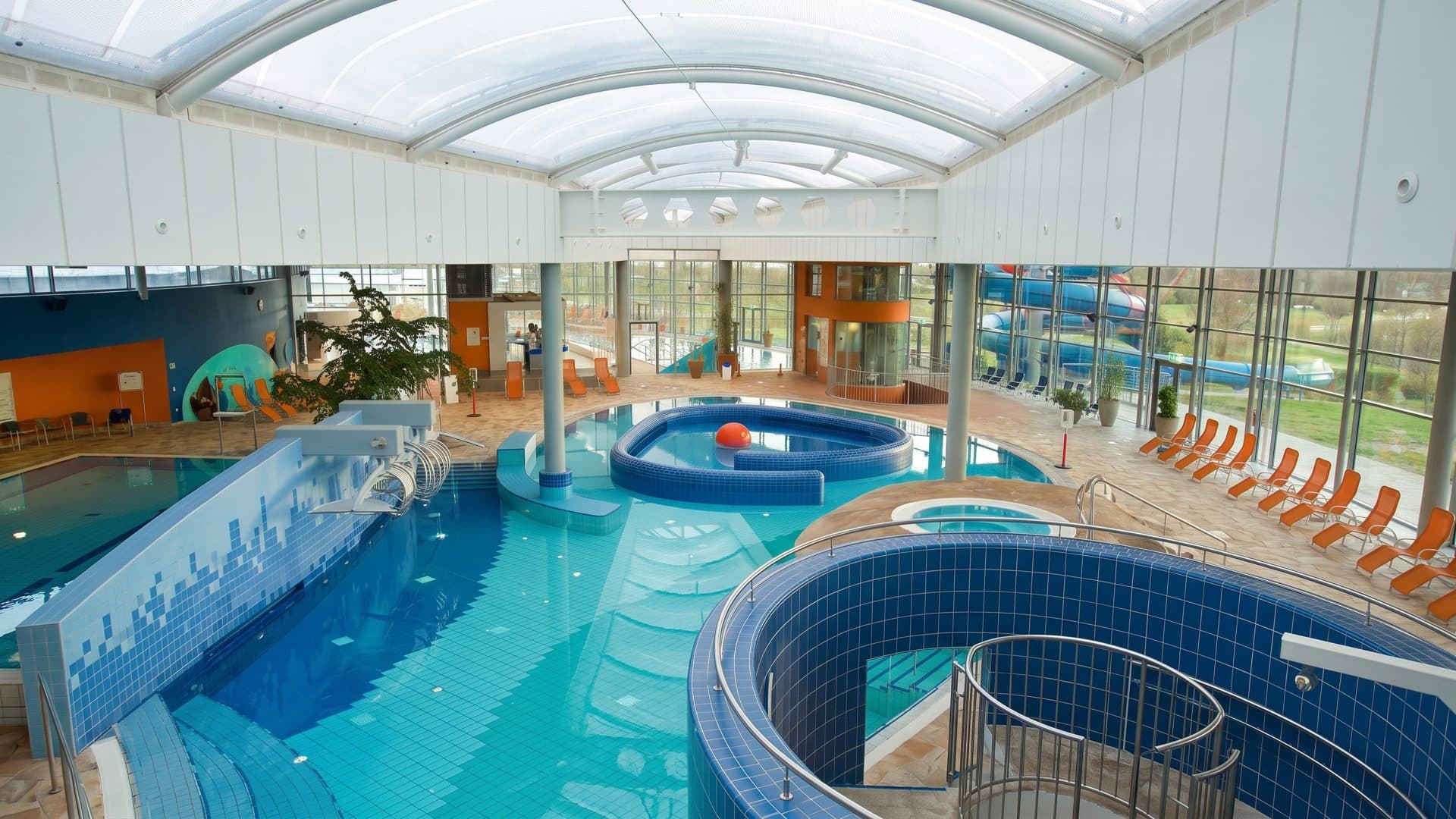Indoor leisure pool under a vaulted roof, with the round whirlpool basin and orange loungers along the glass wall.