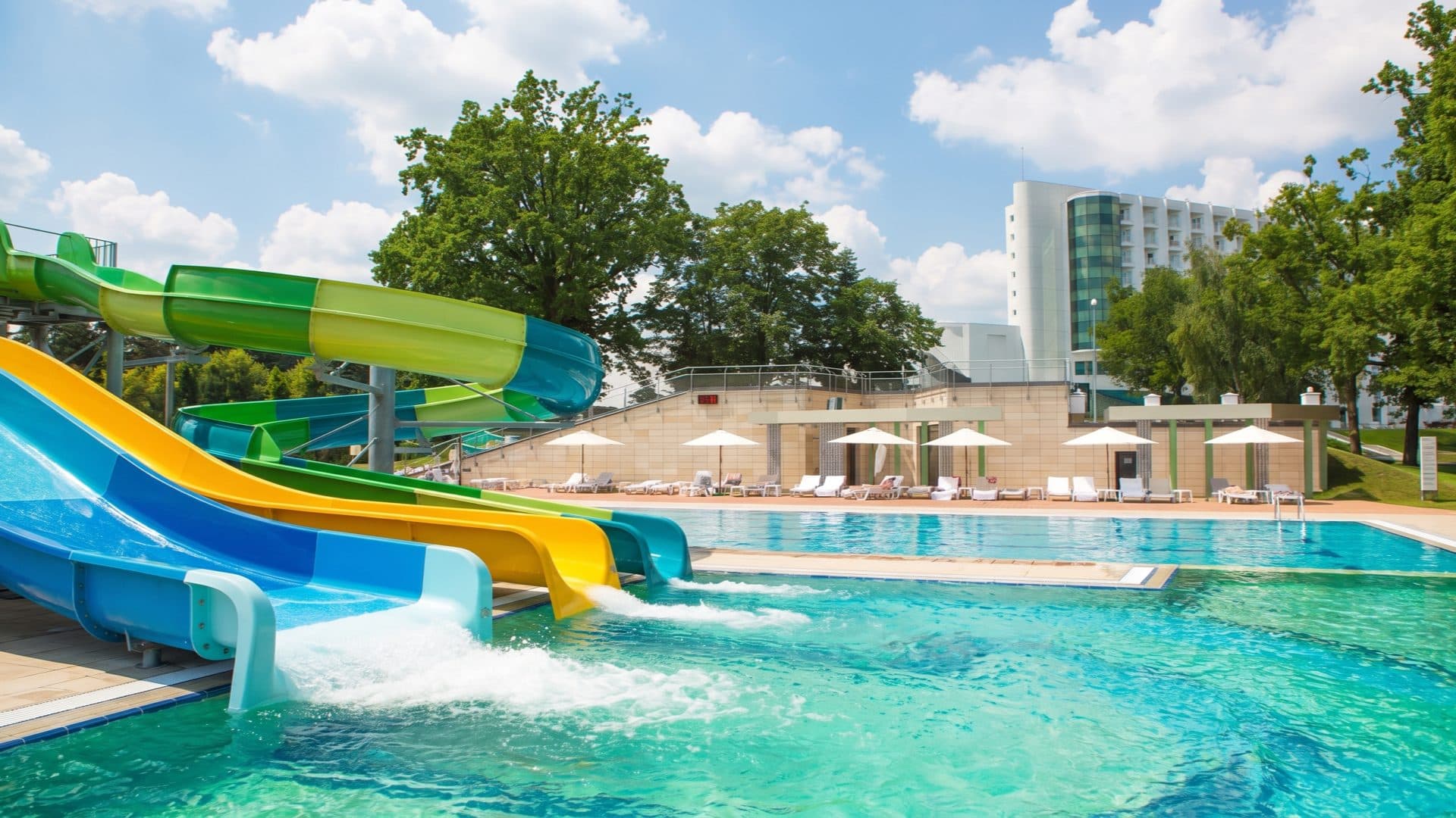 Outdoor resort pool with blue, green and yellow water slides splashing into the water, white parasols and the spa-hotel tower behind.