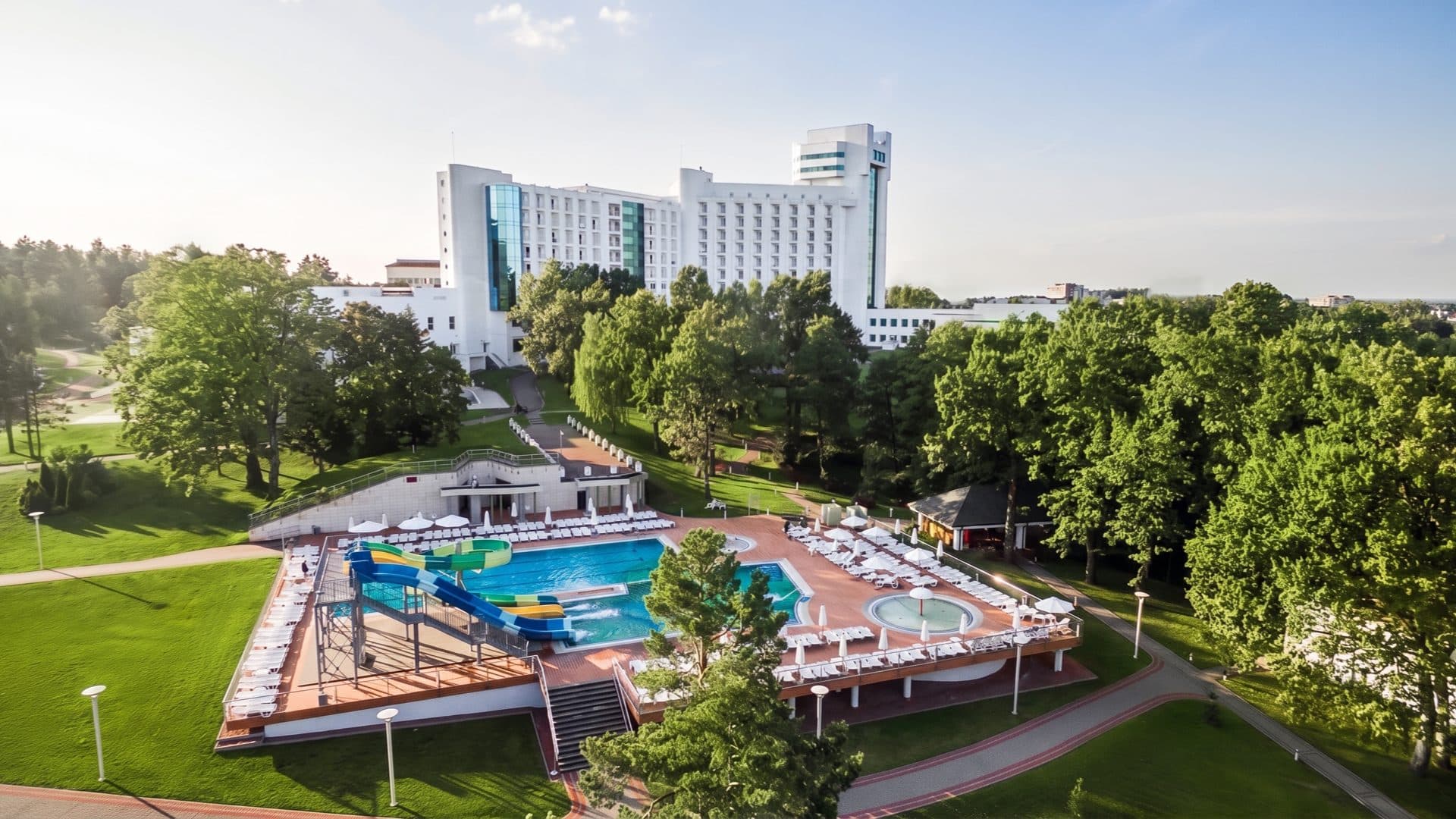 Aerial view of a large white spa-hotel in wooded parkland, with an outdoor pool, water slides and rows of loungers on a terrace below.