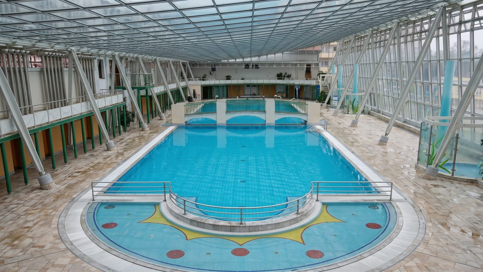 Indoor thermal pool under a glazed steel roof, with a sunburst mosaic on the pool floor