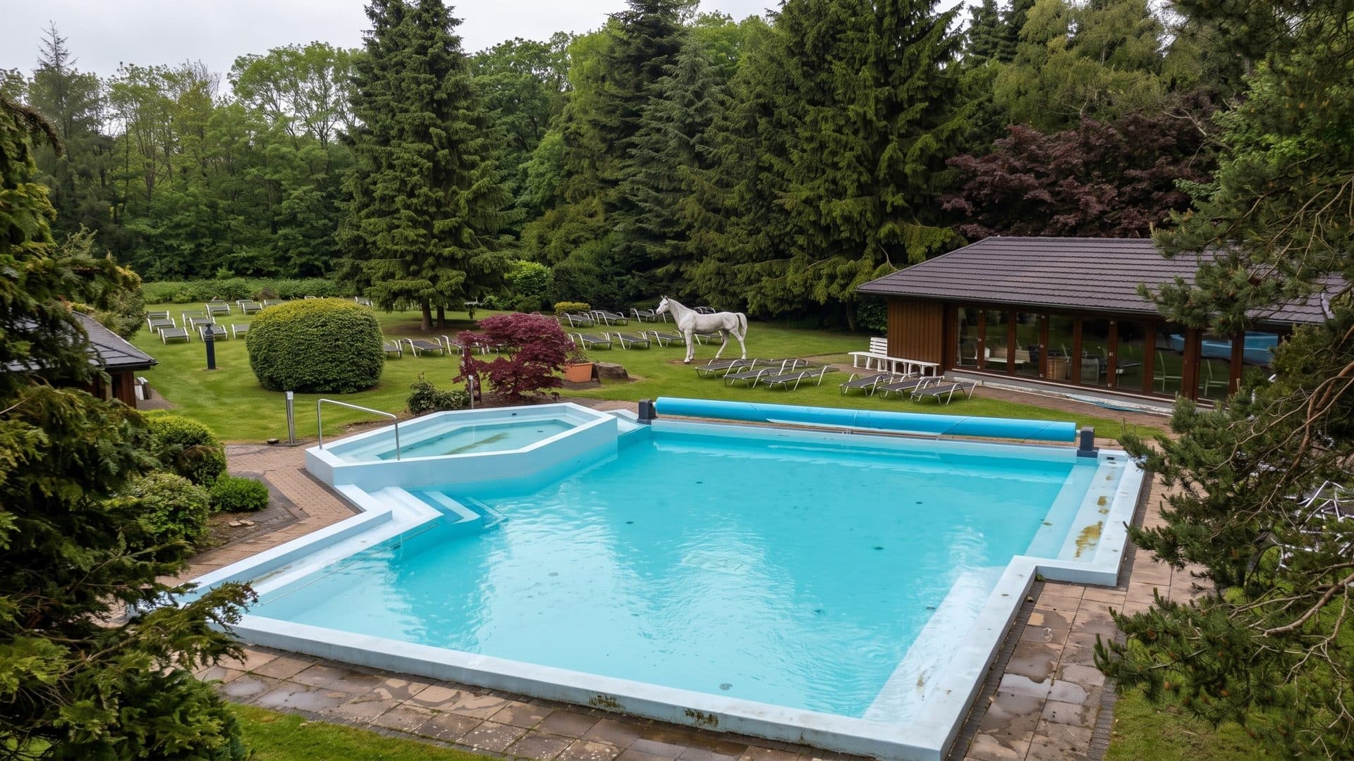 The outdoor pool of the Roetgen Therme in a garden with fir trees, a horse sculpture and a low timber pavilion