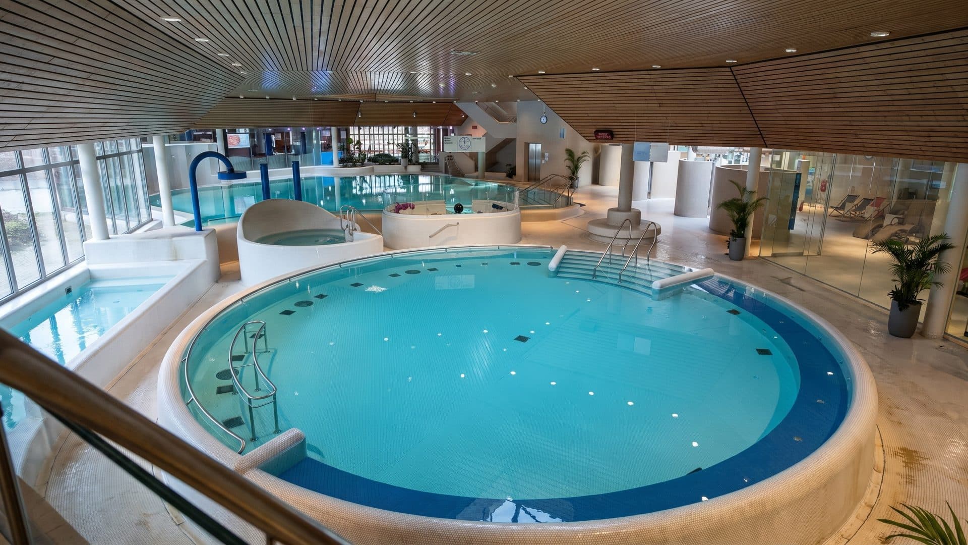 Round indoor thermal pool with a blue rim and a whirlpool, under a timber slatted ceiling