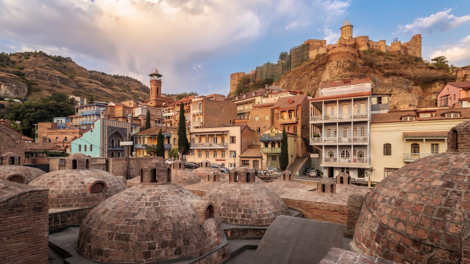 Brick domes of Tbilisi’s sulphur baths below the old town, with a minaret, the hilltop fortress and a church catching the evening light.