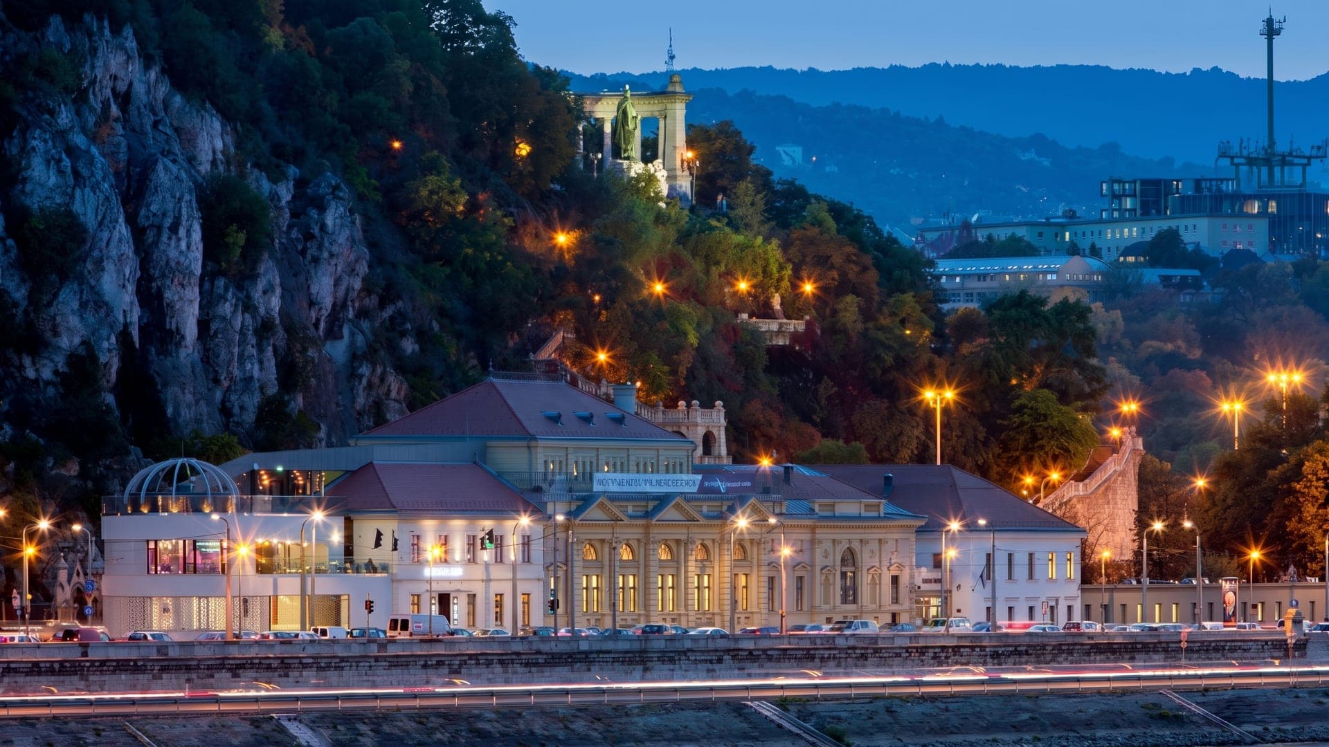 The historic bath lit at night beneath the cliff, with city lights beyond