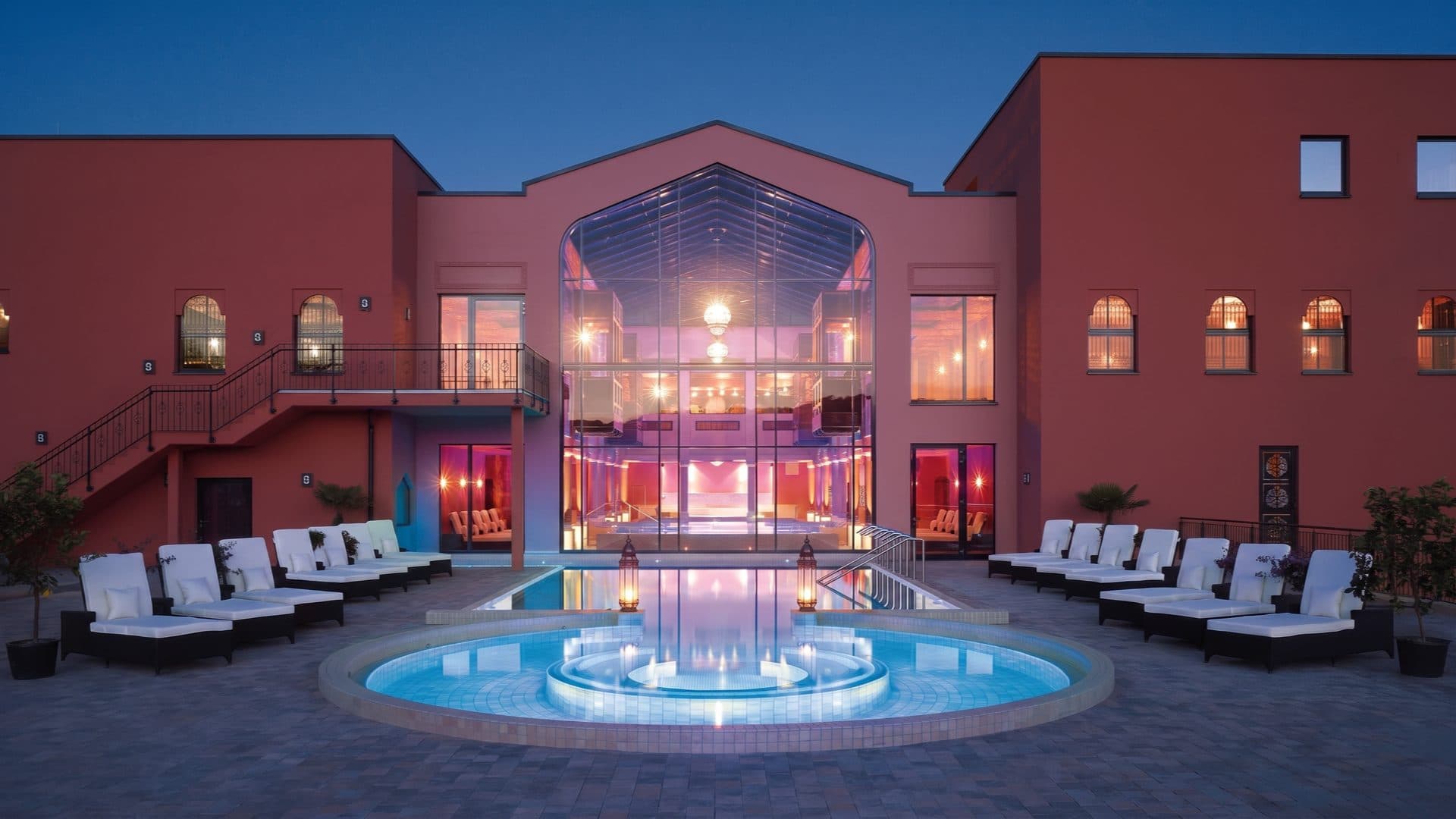 Moroccan-themed spa at dusk, its tall glazed atrium glowing above a round tiled courtyard pool ringed with white loungers and lanterns.