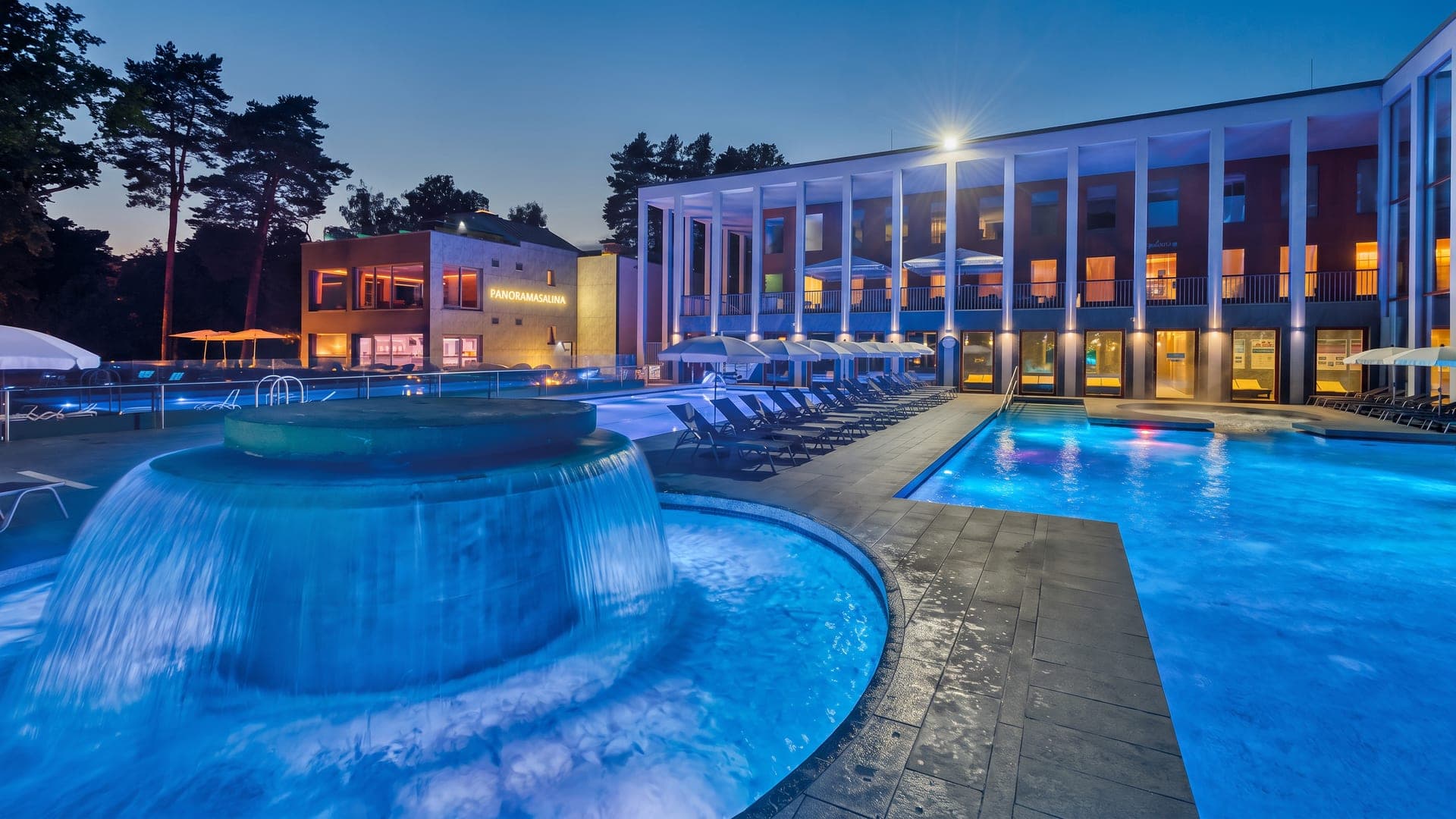 Outdoor pool with a mushroom waterfall lit blue at dusk, beside a colonnaded building