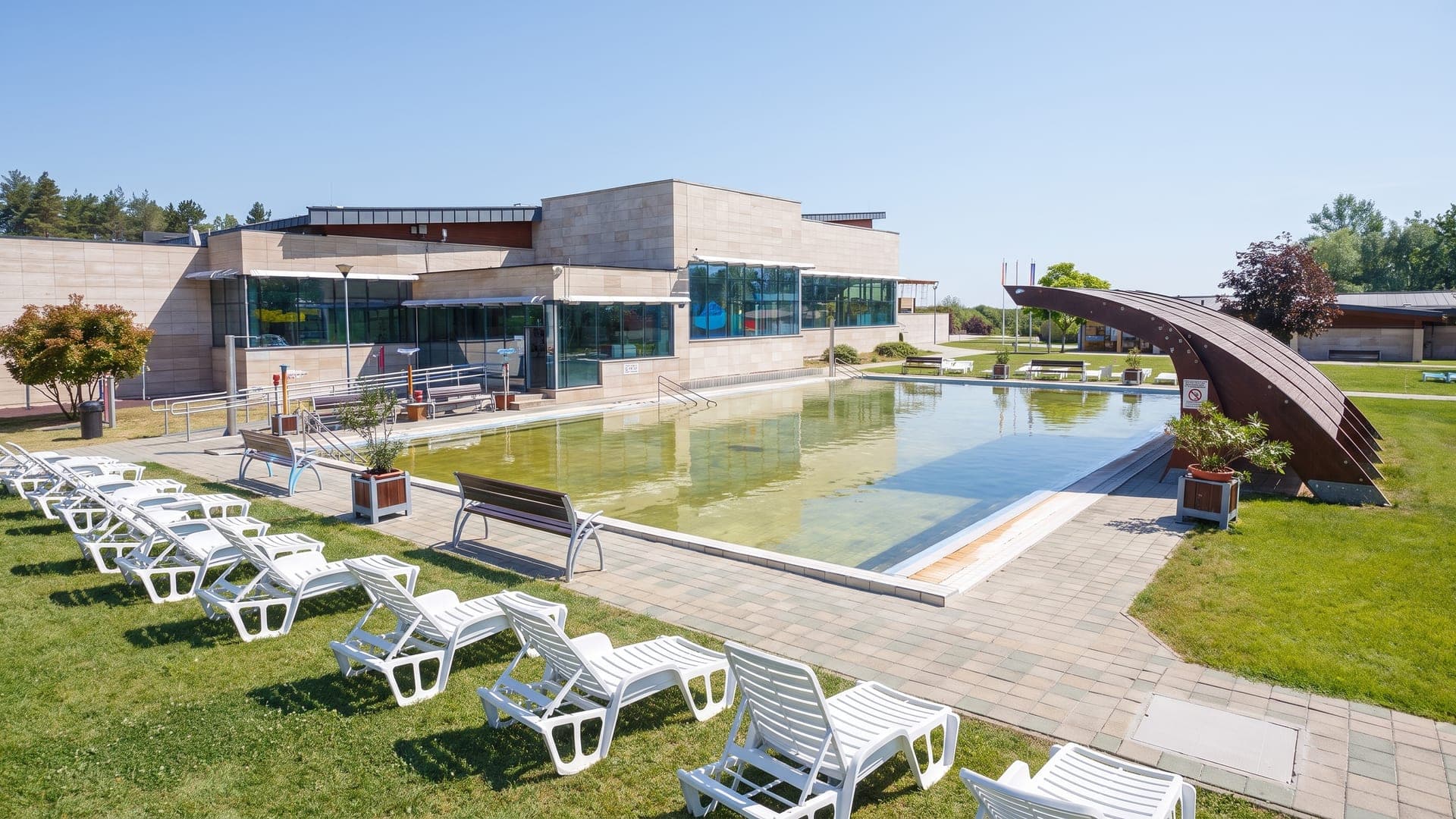 The outdoor pool at the Sarkany bath in Nyirbator, in front of the low concrete-and-glass building with a dragon sculpture beside it.
