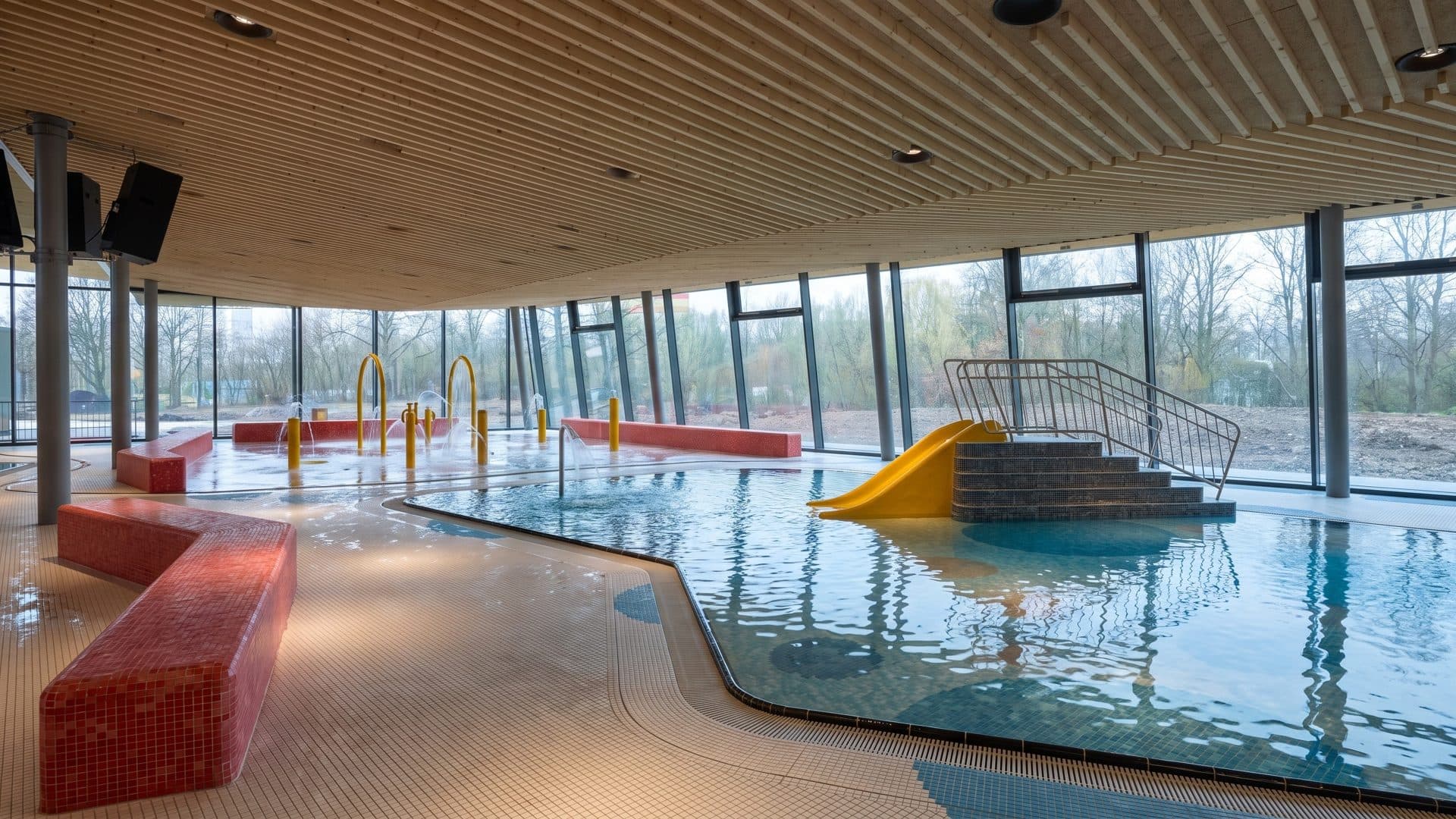 Indoor children's pool with a yellow slide, red tiled benches and a timber ceiling, seen through tall windows