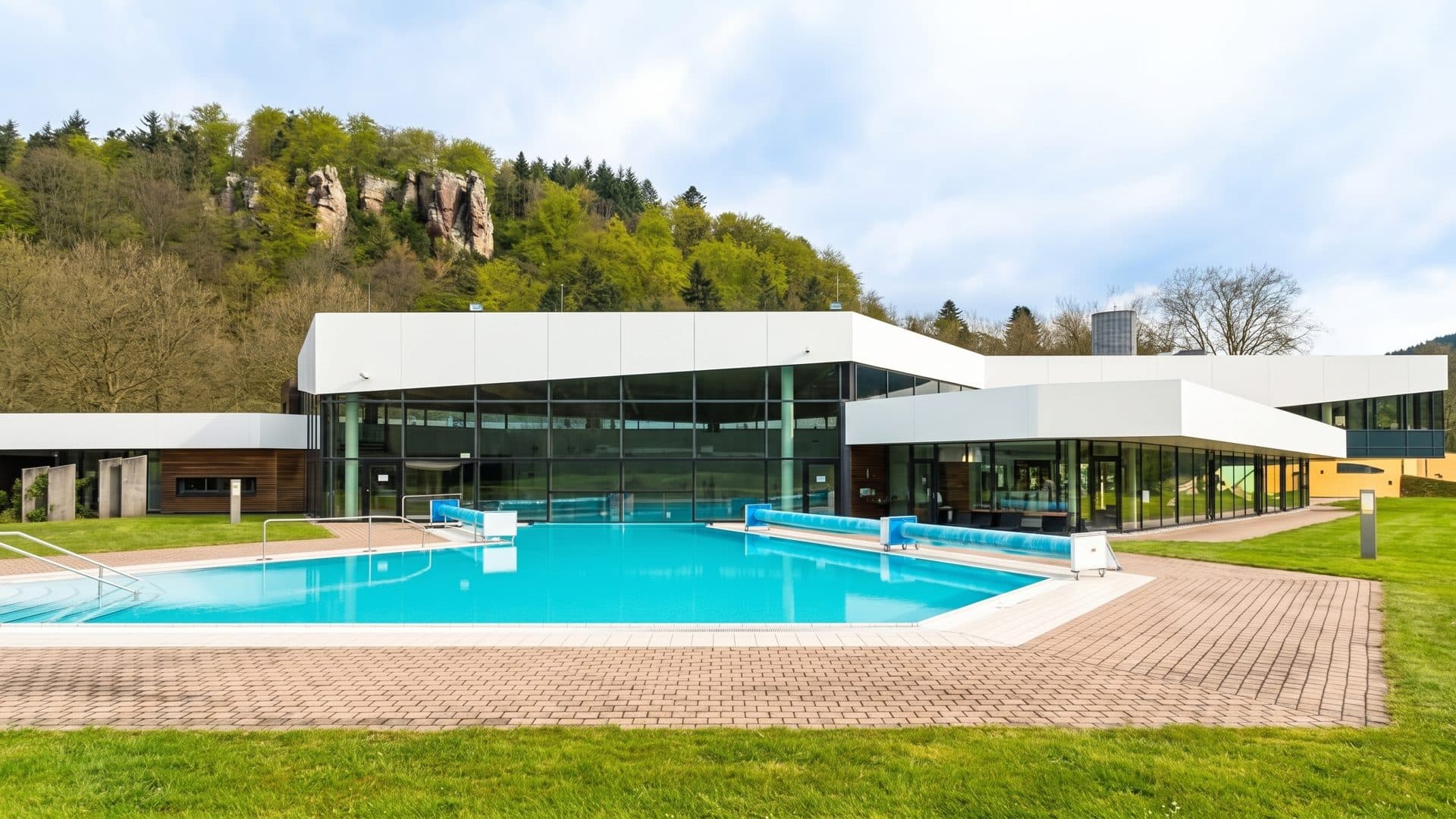 Low glass building of the baths with its outdoor pool in front, wooded hills and cliffs behind.