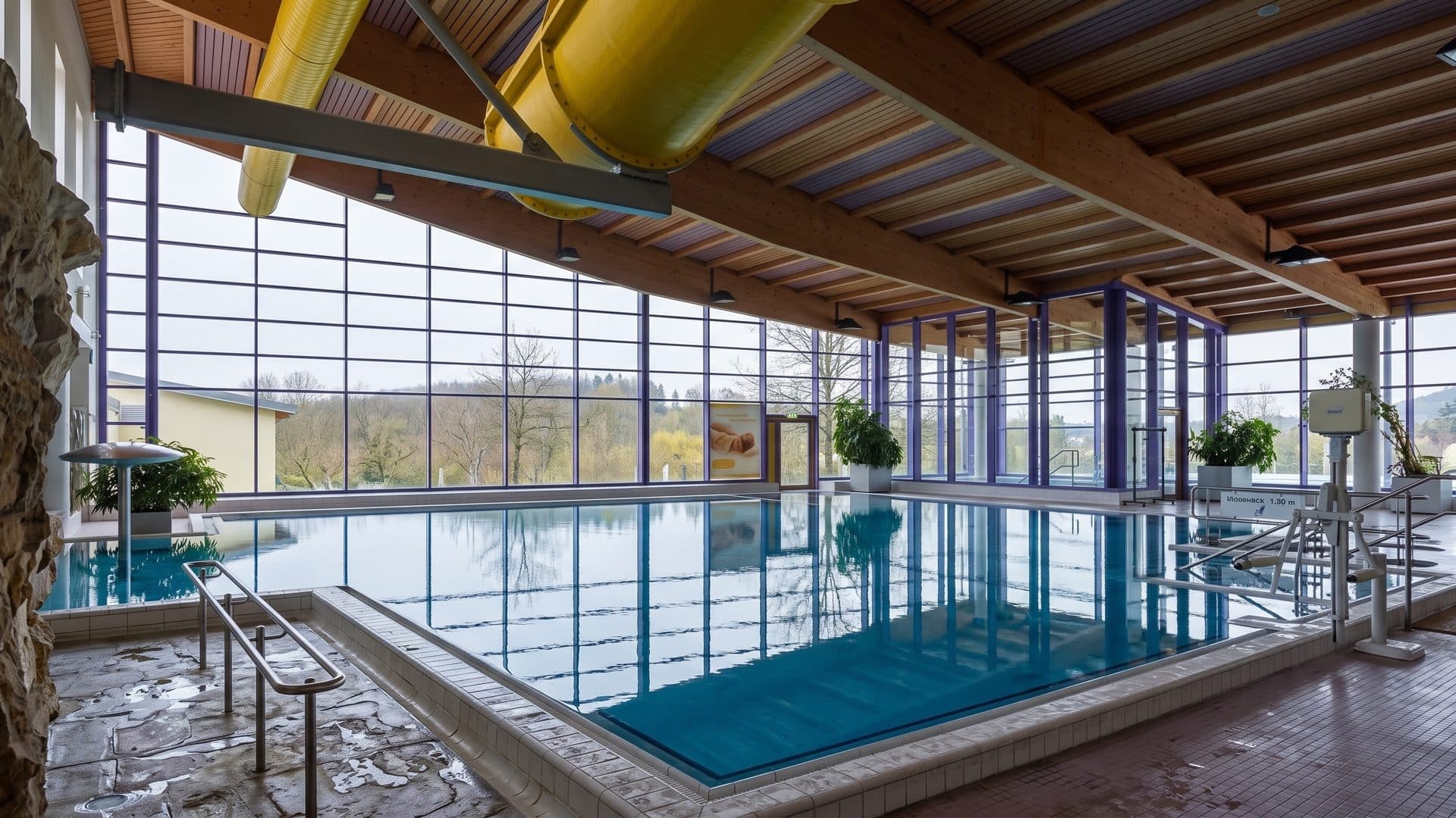 The indoor pool of the Silber-Therme in Warmbad under a timber roof, a yellow slide and a glass wall