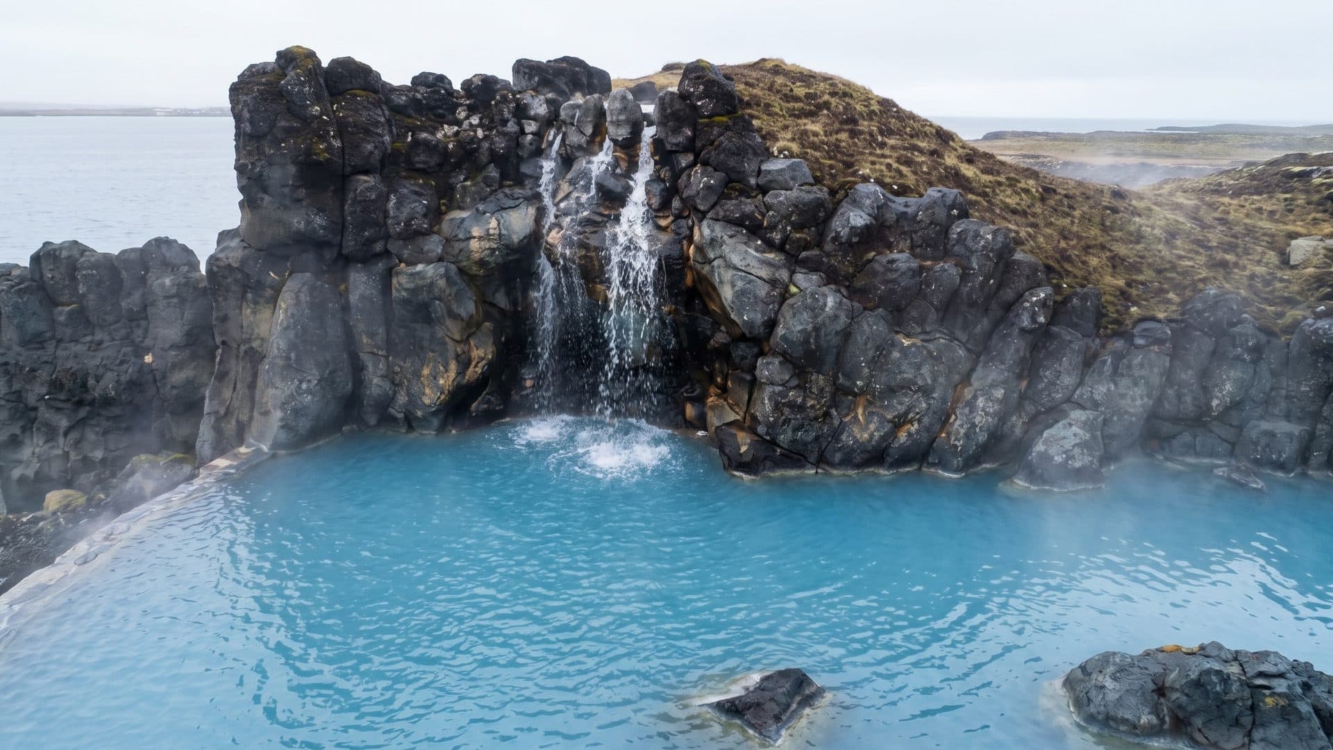 Blue geothermal pool at the foot of a dark basalt cliff with a small waterfall, the sea beyond