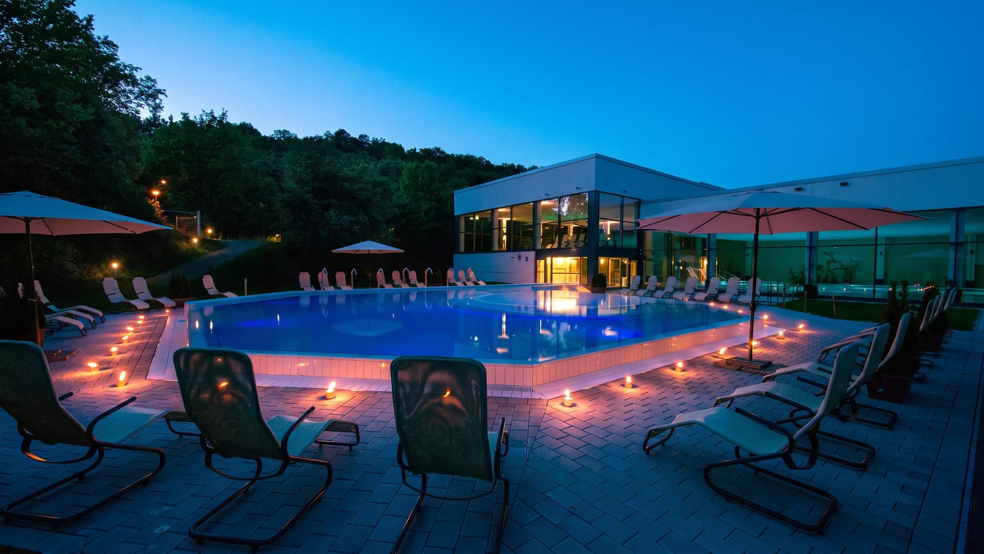 Outdoor pool lit at dusk, with loungers and parasols beside the glass building