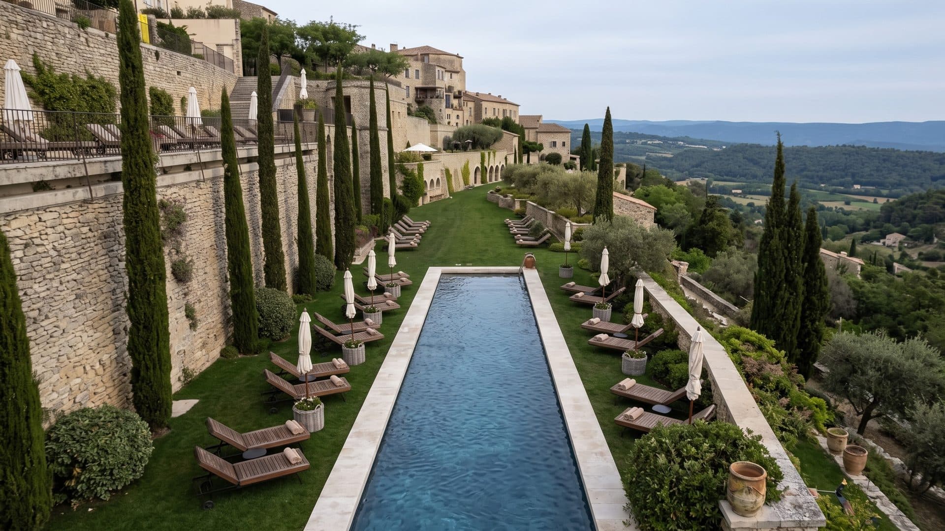 A long narrow pool on a terrace below stone walls and cypresses in Gordes, the Luberon valley beyond