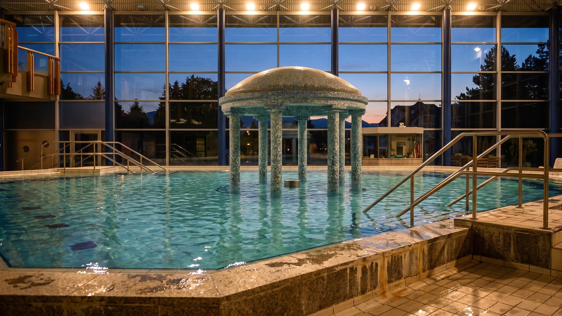 The colonnaded rotunda standing in the thermal pool at the Spessart-Therme, with the glass front and the evening sky behind it