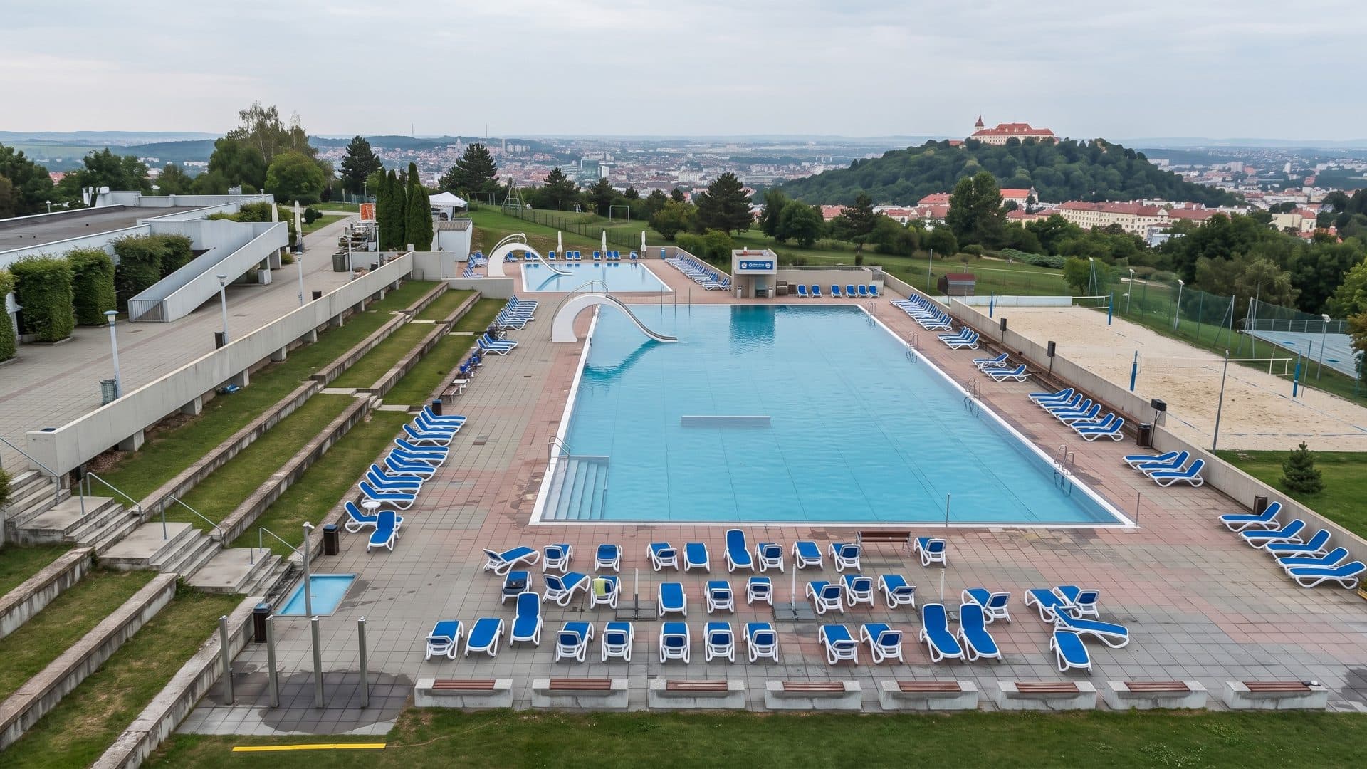The long outdoor pool of the Kravi hora area in Brno with rows of loungers, Spilberk castle on the hill beyond