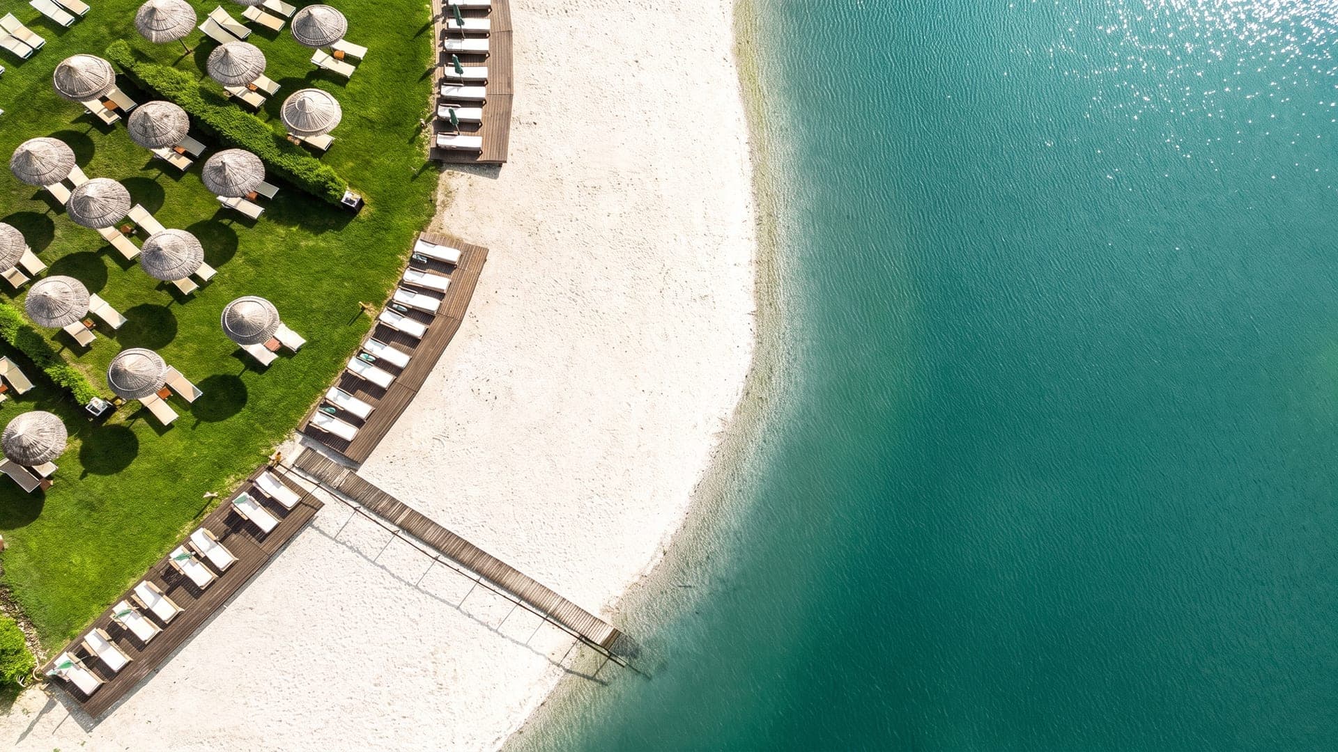 Sandy edge of the turquoise lagoon from above, with loungers and straw parasols on the lawn