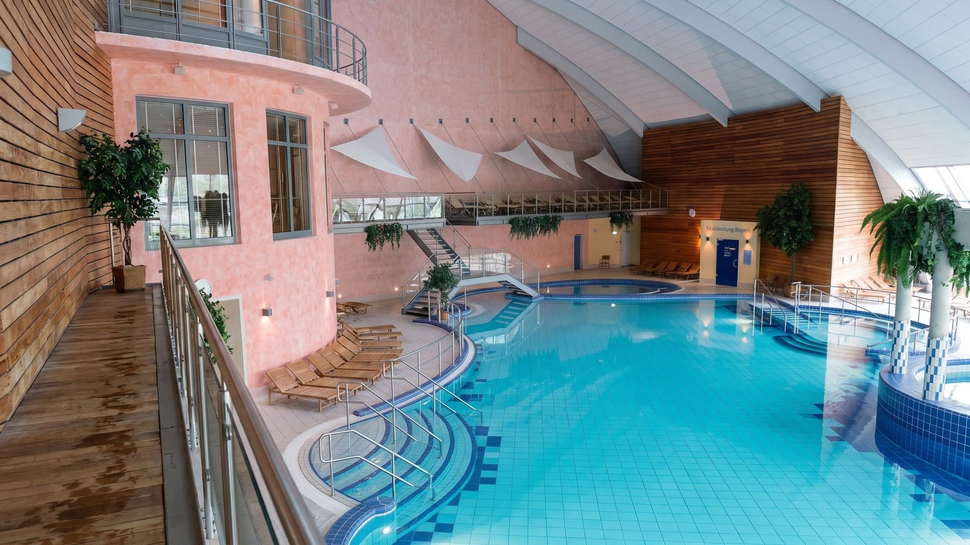 Indoor thermal pool with curved steps below a pink tower and sail-shaped ceiling panels