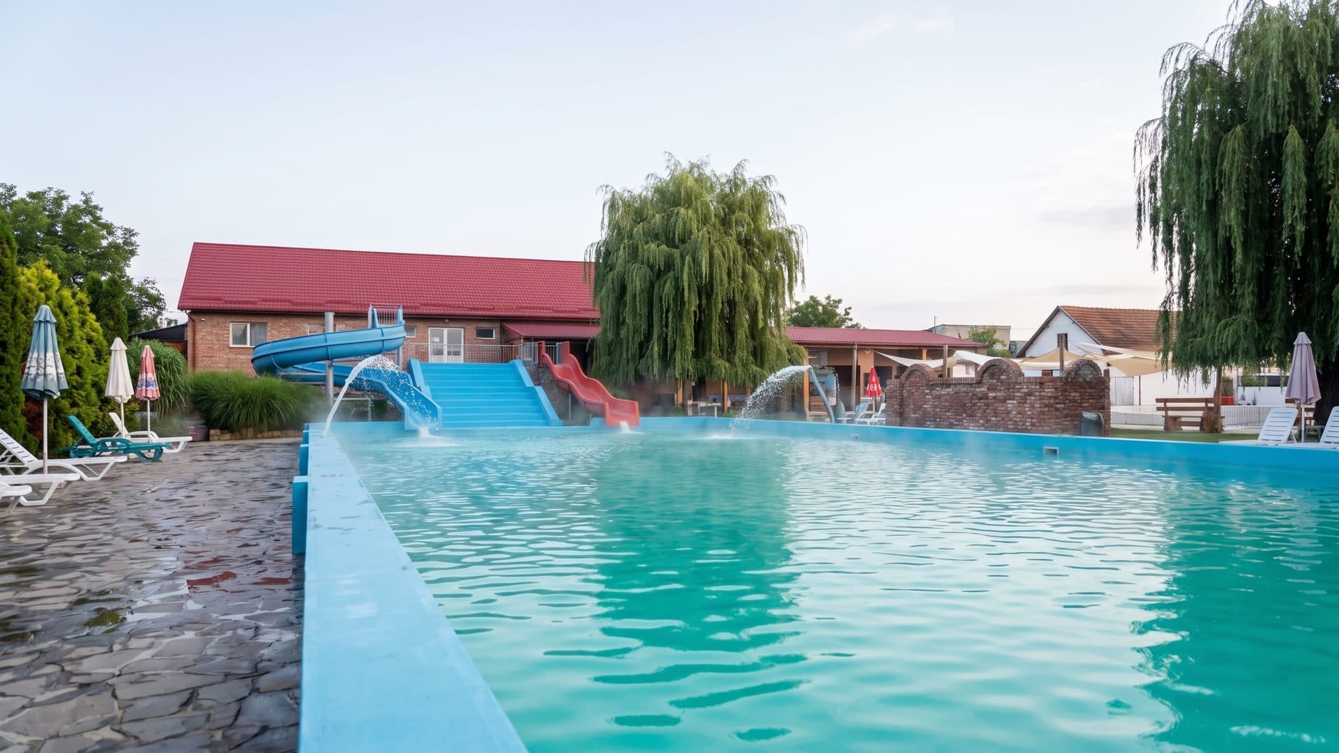 The large outdoor thermal pool at Carei full of clear water, willows and brick farm buildings behind it and loungers along the paved edge