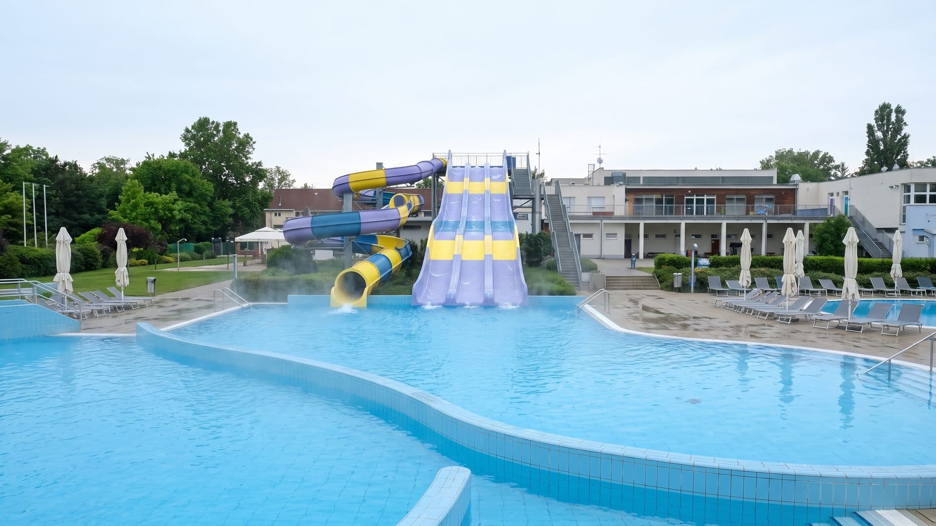 The outdoor pool at Strandul Neptun with a slide tower at one end, parasols along the deck and the bathhouse behind