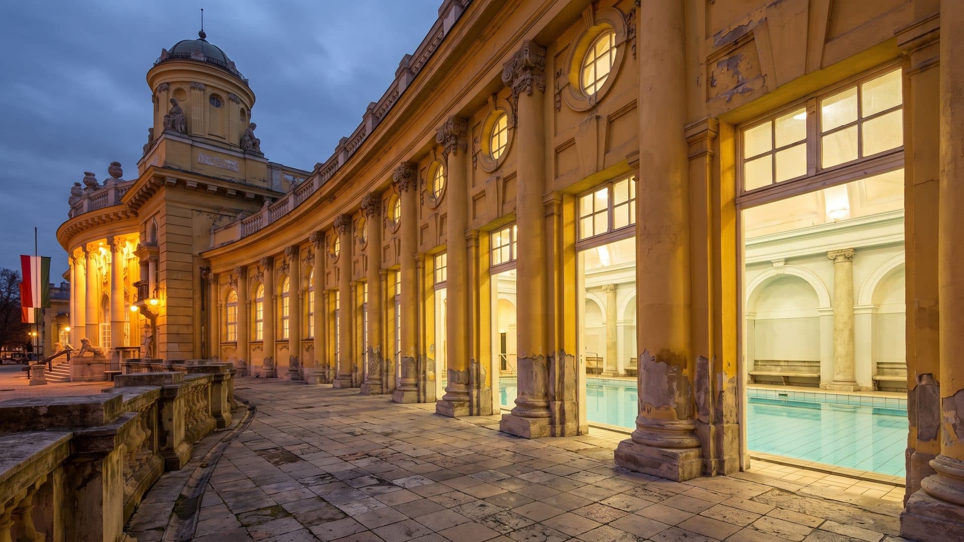 The colonnaded courtyard of the Szechenyi baths at dusk, its yellow arcades lit above the outdoor pool