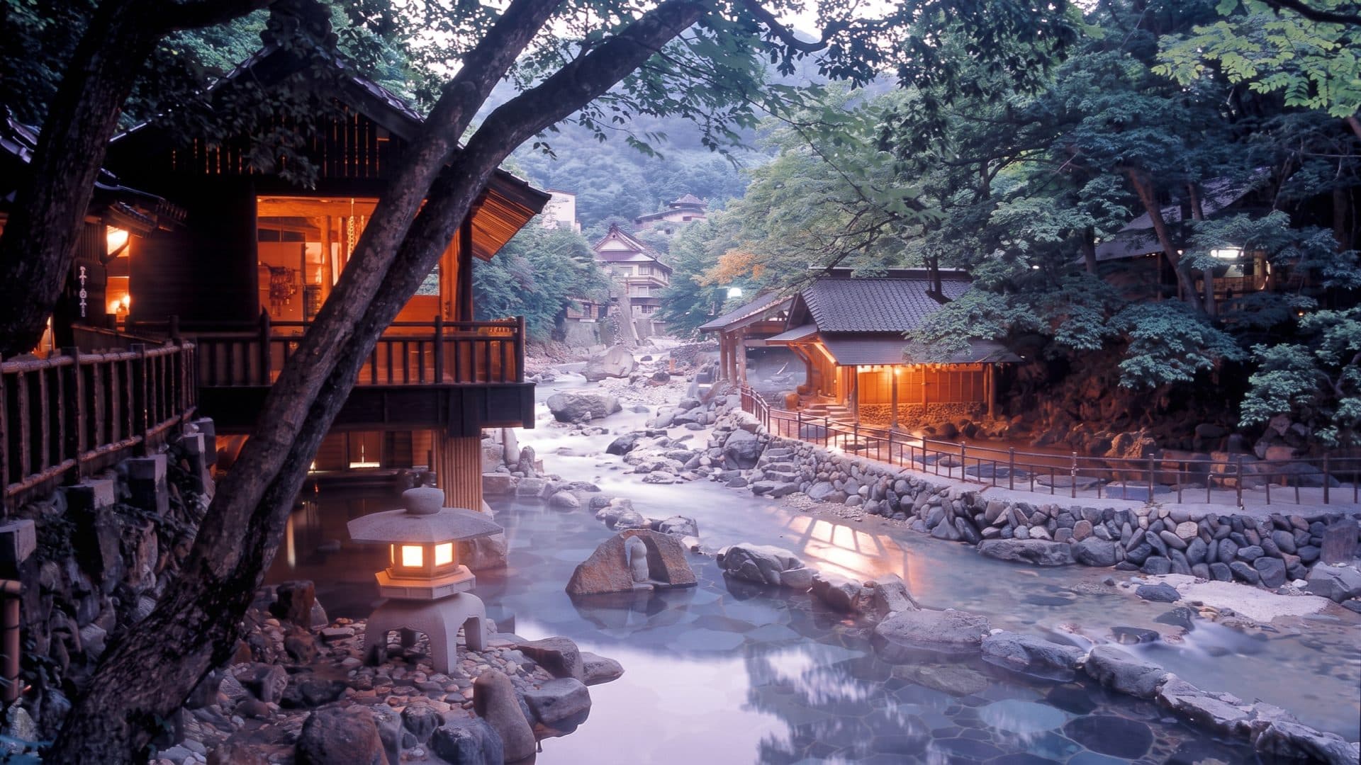 Riverside onsen at dusk, timber ryokan buildings lit above milky rock-lined bathing pools and a stone lantern by the water.