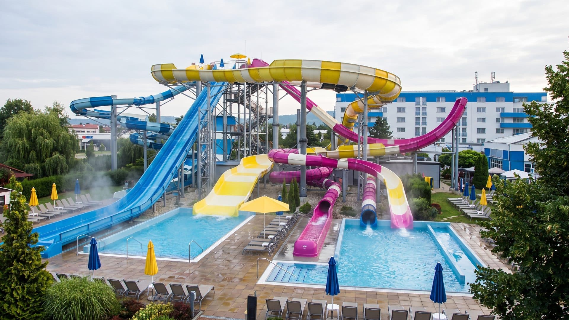 The slide park at Tatralandia, a bundle of yellow, pink and blue flumes above the outdoor pools with the mountains behind