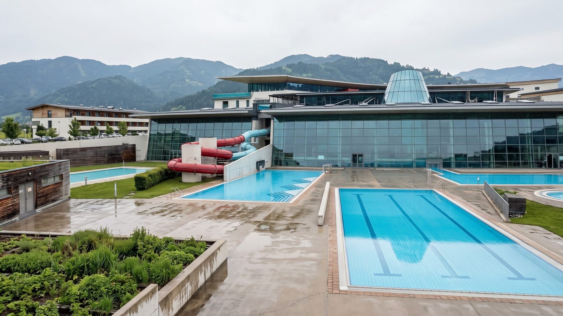 Outdoor pools of the Tauern Spa Kaprun in front of the glass bath hall with its slides, mountains behind