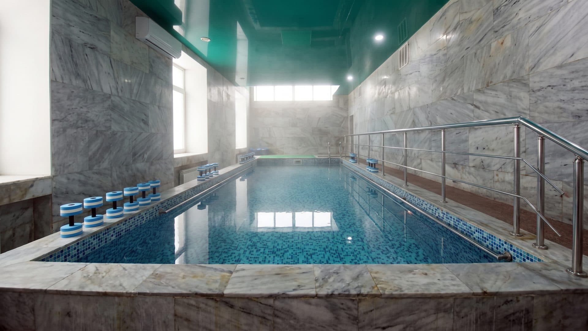 Marble-lined indoor pool hall of the balneological resort, a handrail down one side and aqua-fitness floats stacked at the edge.
