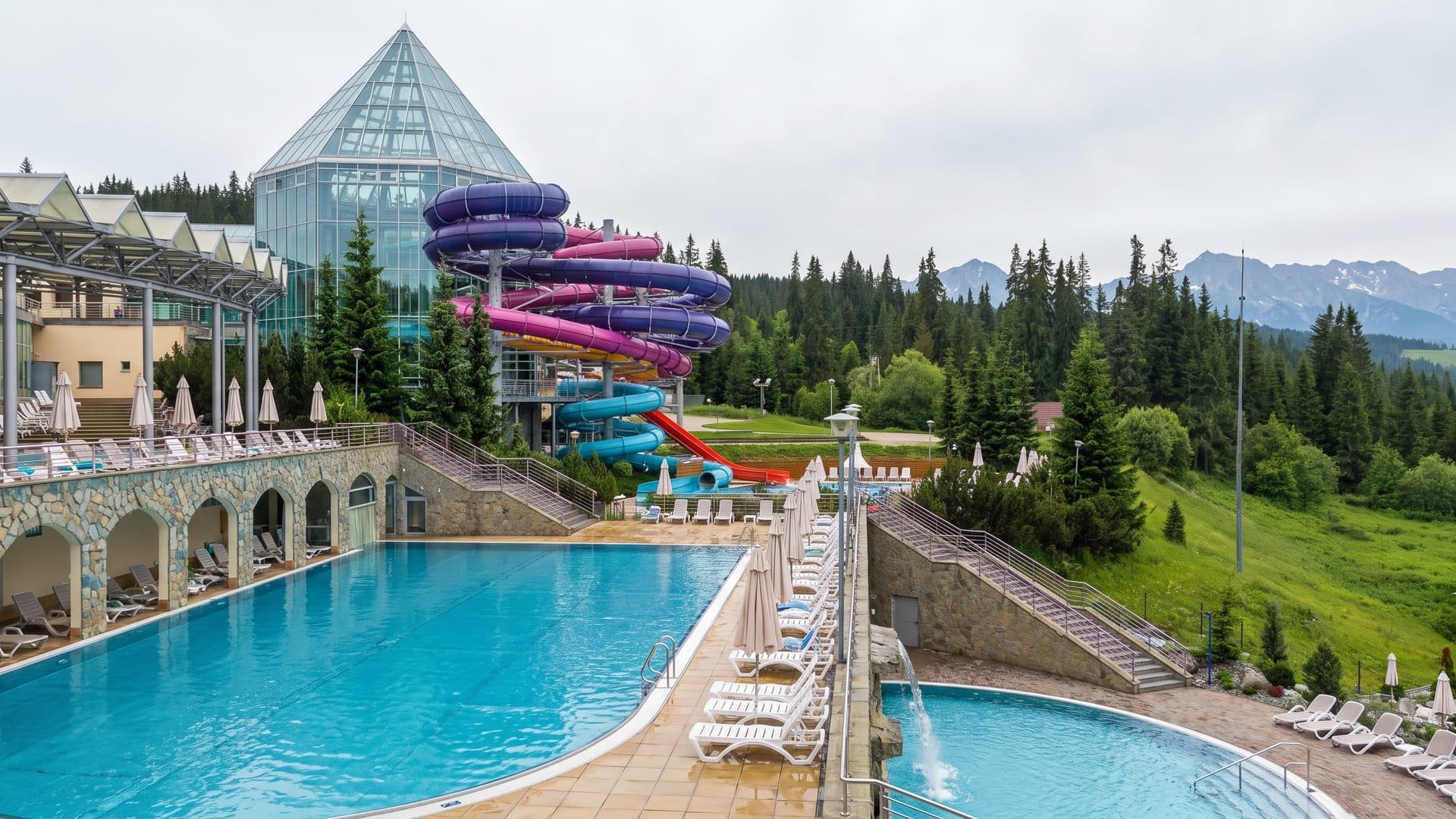 The outdoor thermal pool at Terma Bukovina below its glazed hall and slide tower, with the Tatra meadows and forest beyond