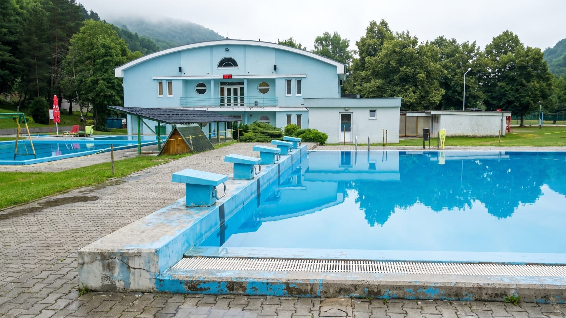 Outdoor swimming pool with starting blocks, the pale blue bath building and wooded hills behind