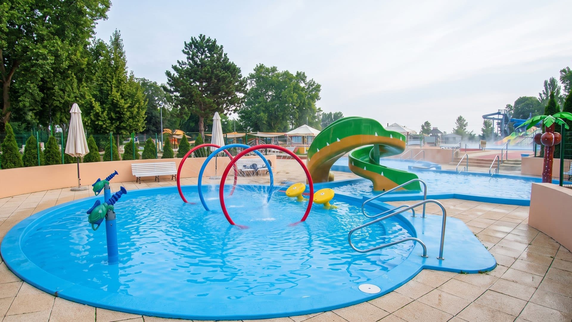 The children's splash pool at the Emil Tatarik baths with a green frog slide and water arches, parasols on the paving around it