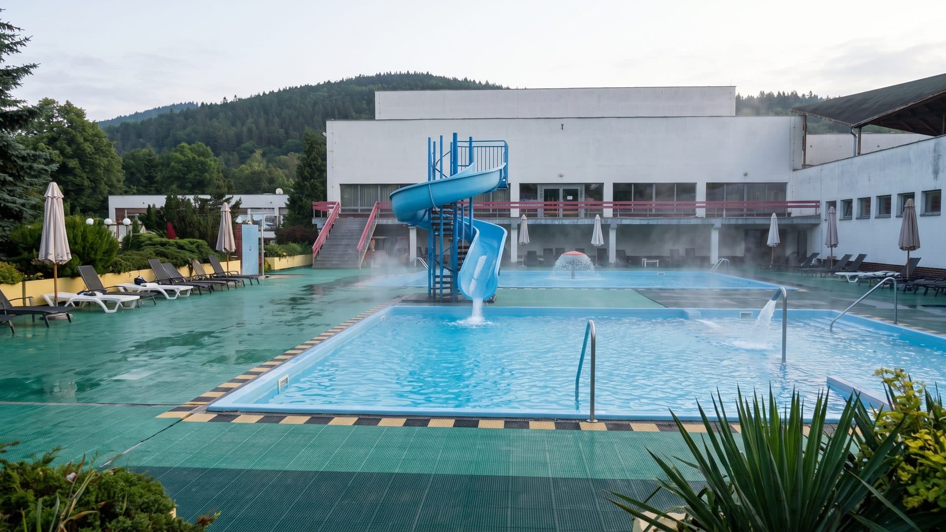 The outdoor thermal pool at Veronika in Rajec steaming below the bathhouse, a blue slide at one end and wooded hills behind