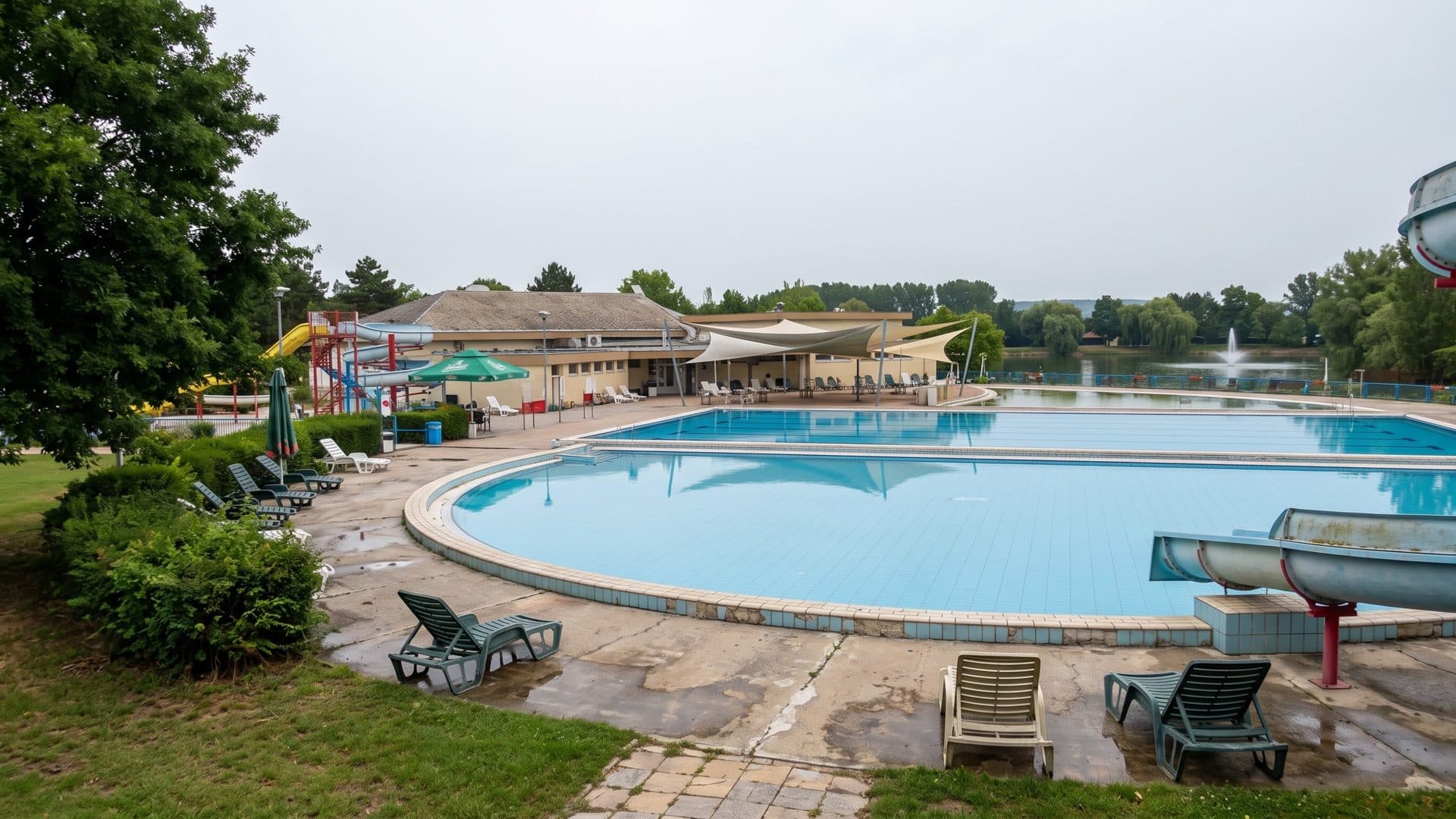 The round open-air pool of the Vincov les thermal lido with loungers on the deck, a slide at the edge and the lake beyond