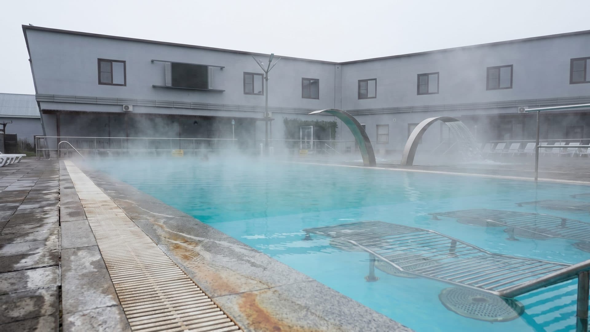 A steaming milky-blue thermal pool at Psygups alongside the long low bathhouse, its deck crusted with minerals