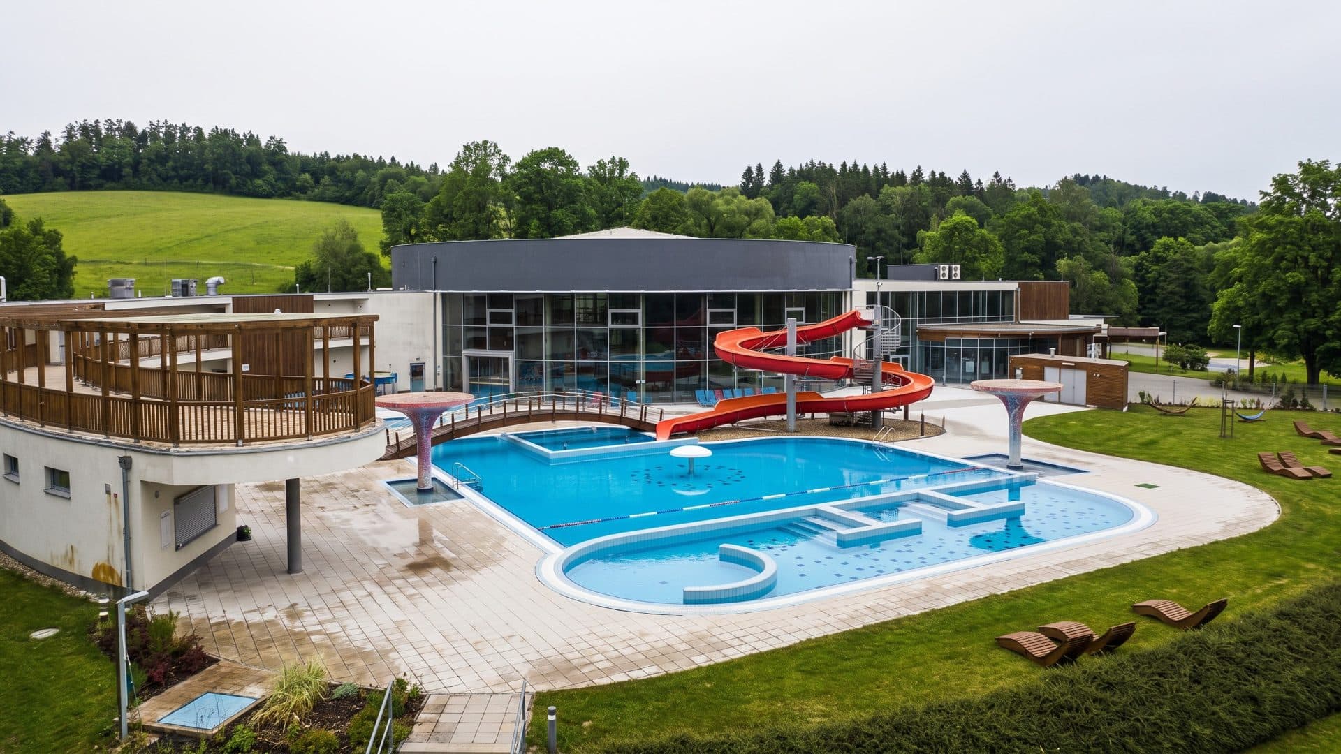 Outdoor thermal pool with a red spiral slide beside the glazed pool hall, green hills behind