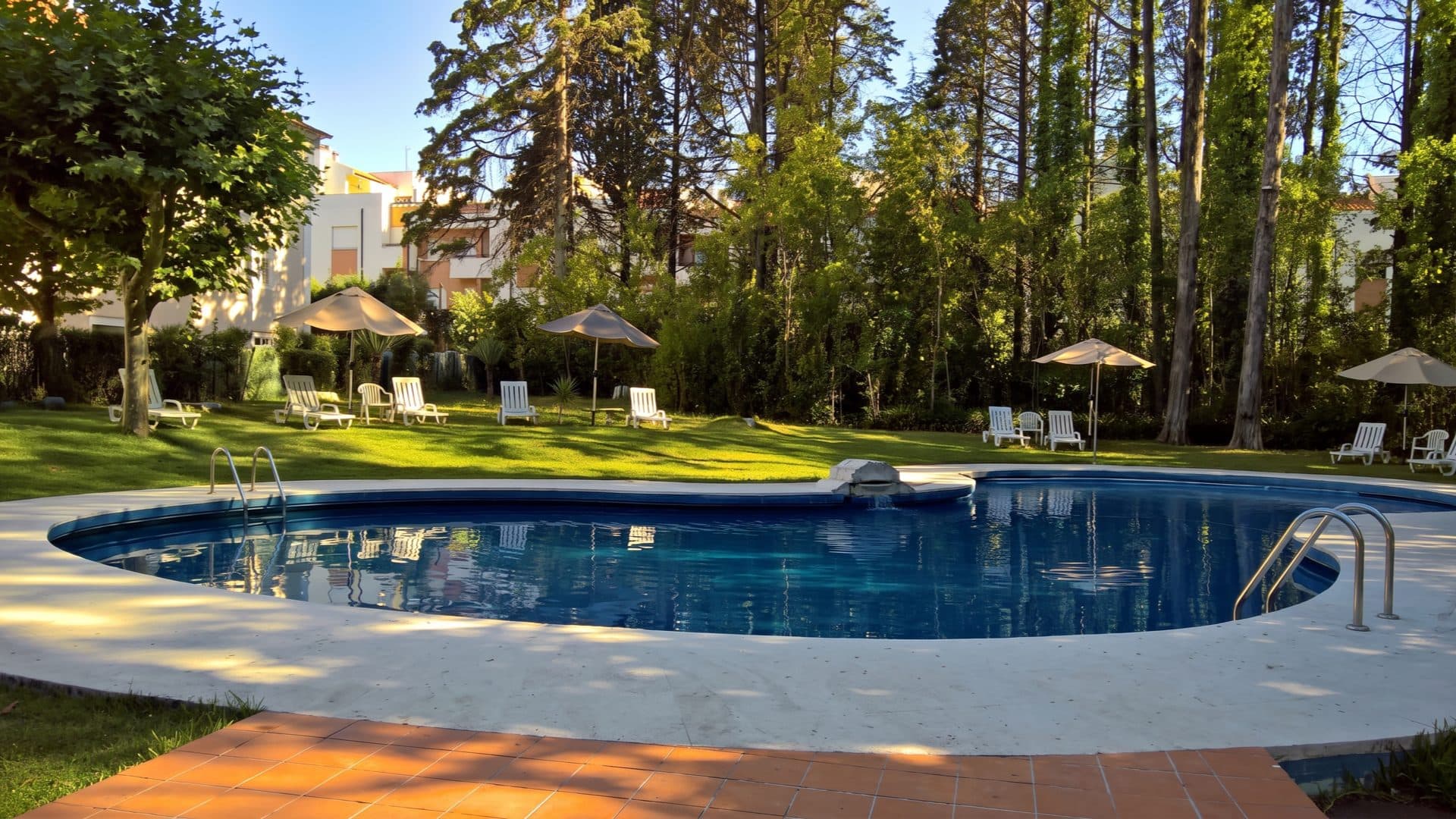 Oval garden pool in a wooded spa park at golden hour, white loungers and parasols set on the surrounding lawn.