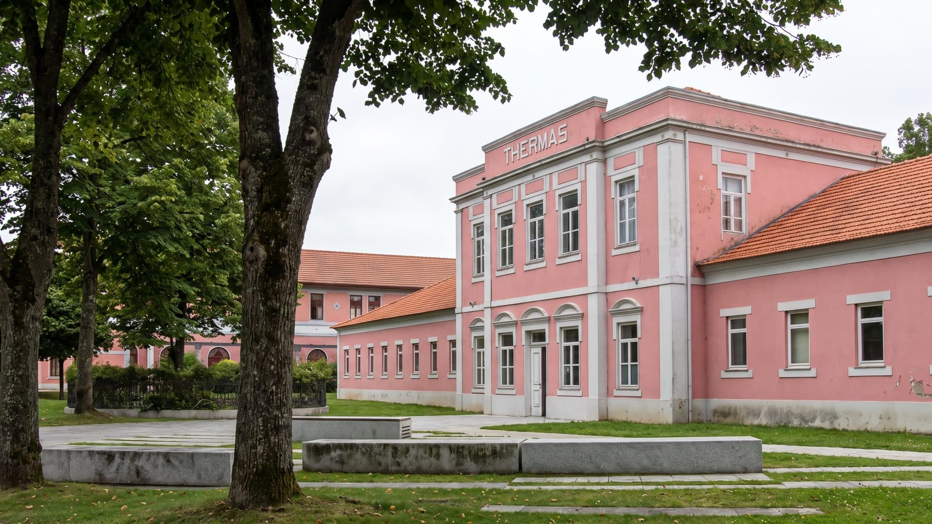The salmon-pink bath house of Caldas das Taipas seen across its lawn between mature trees