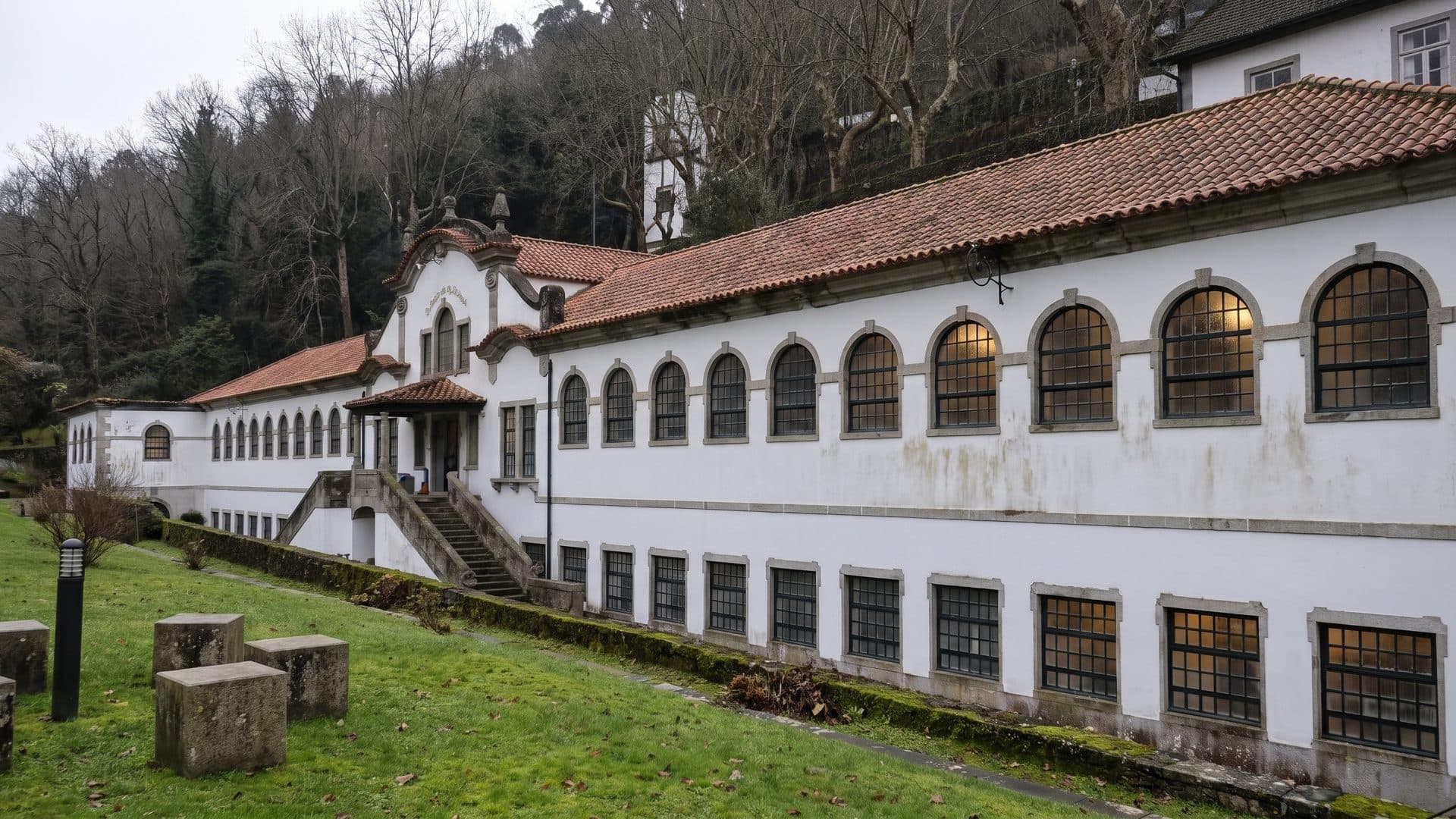 The long white arcaded bath house of Termas de Caldelas on its lawn below a wooded hillside