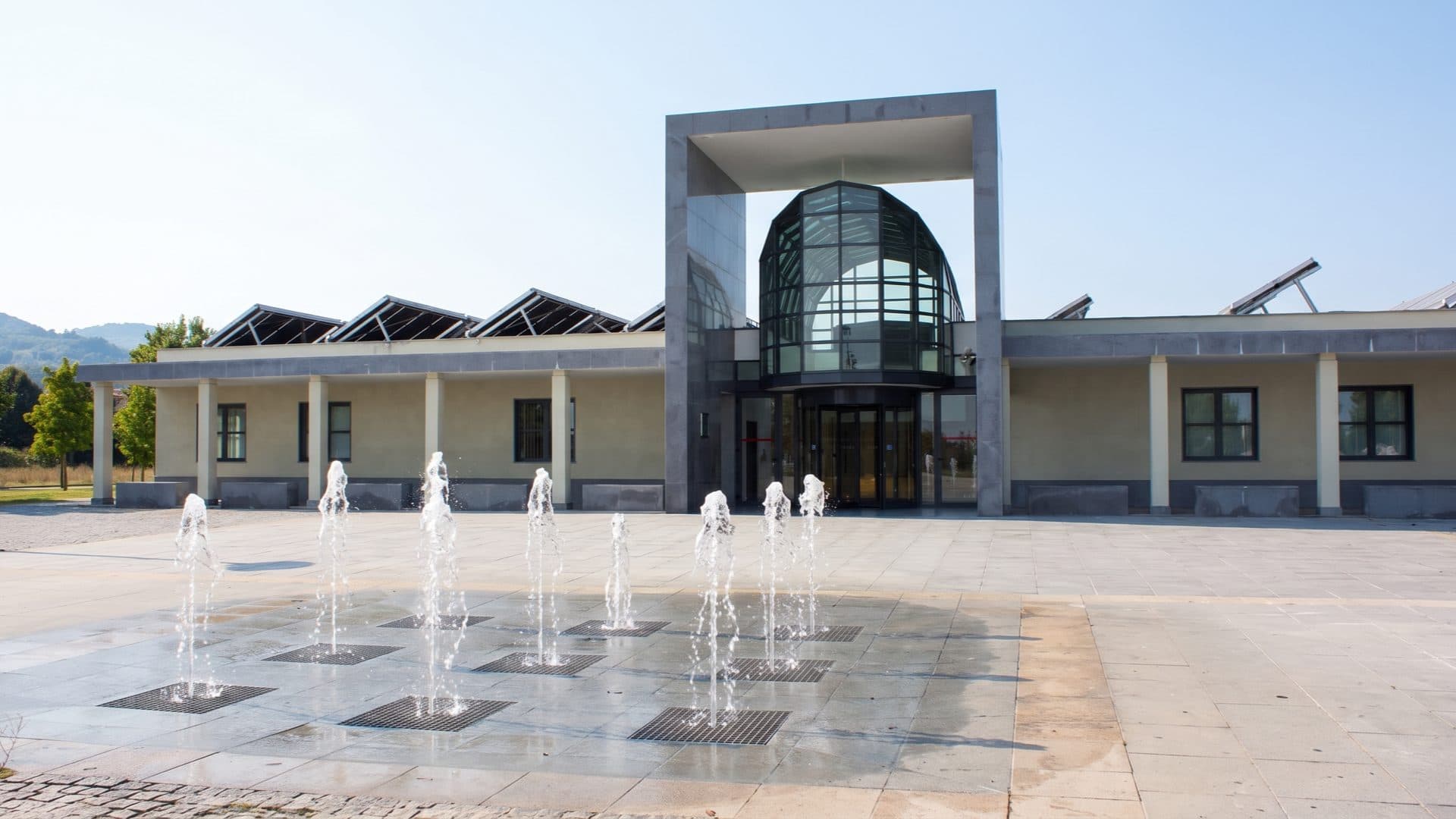 Modern spa building with a glazed rotunda entrance, jets of a ground fountain playing across the forecourt.