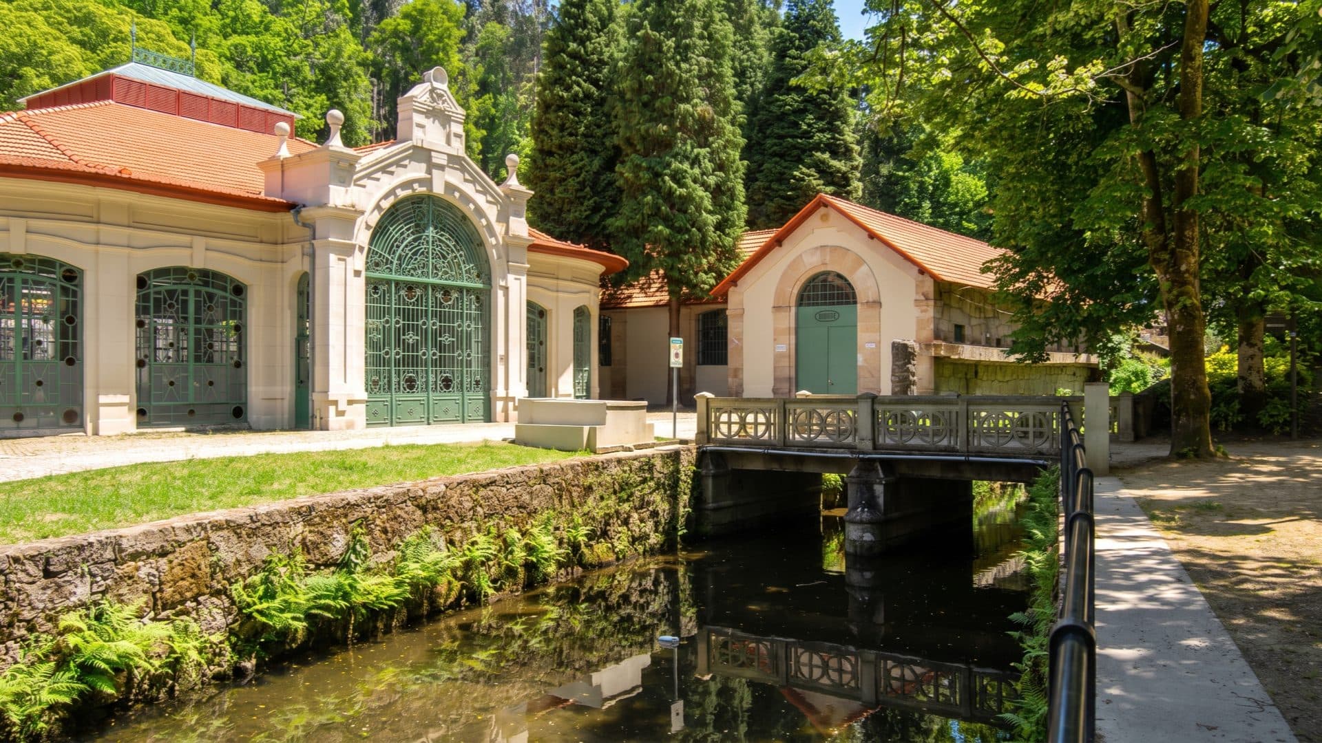 Art Nouveau spa pavilions with an ornate green iron-and-glass portal and a small pump house, a stone footbridge crossing a fern-lined stream.