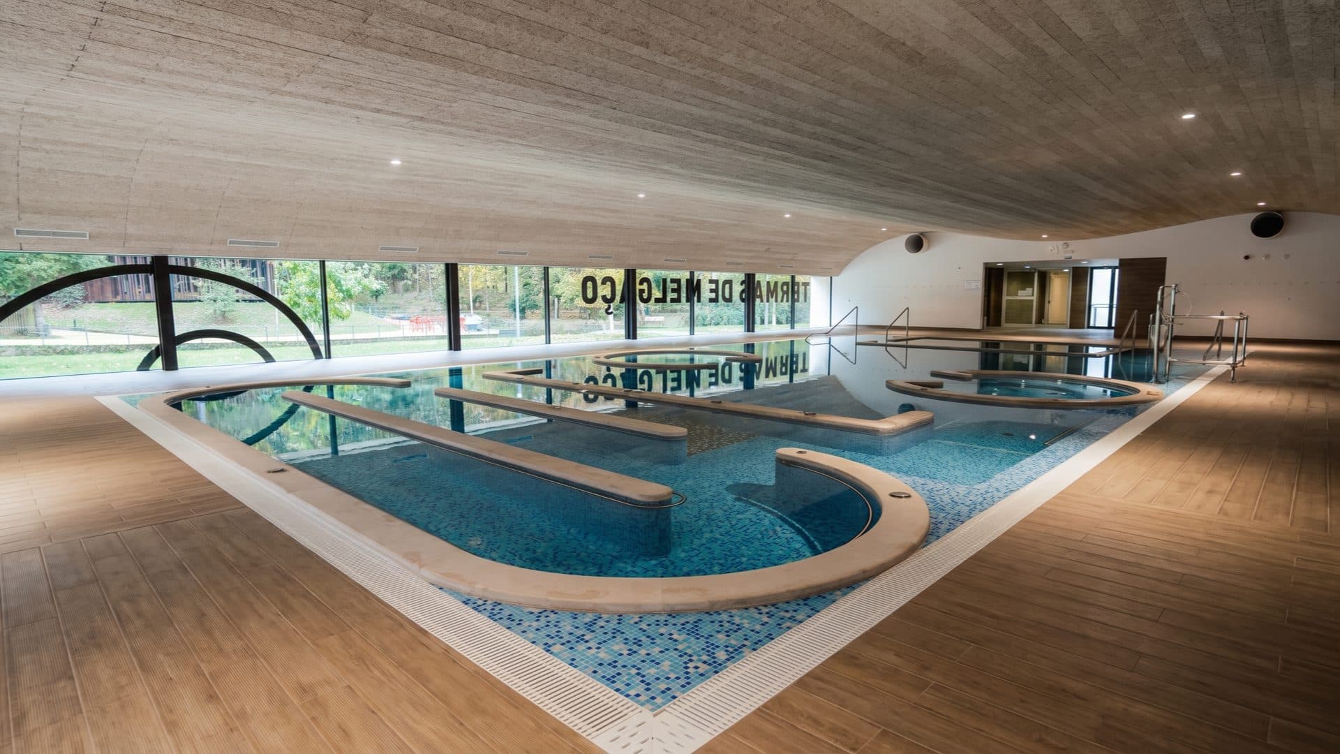 Modern indoor thermal pool hall with curved pool forms and shaped island seats under a wave-form timber ceiling, floor-to-ceiling glass onto the park.