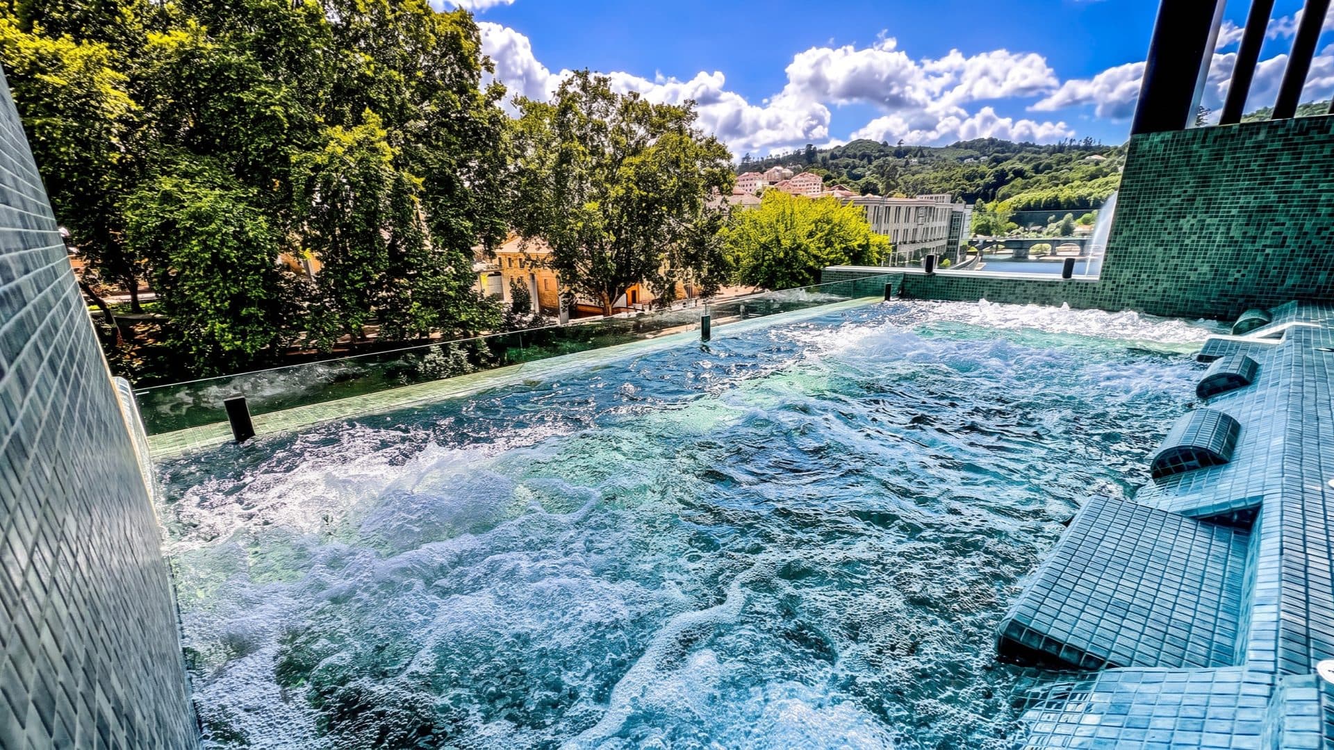 Rooftop hydro pool in green mosaic tile, water bubbling against a glass balustrade above a river valley and hillside town.