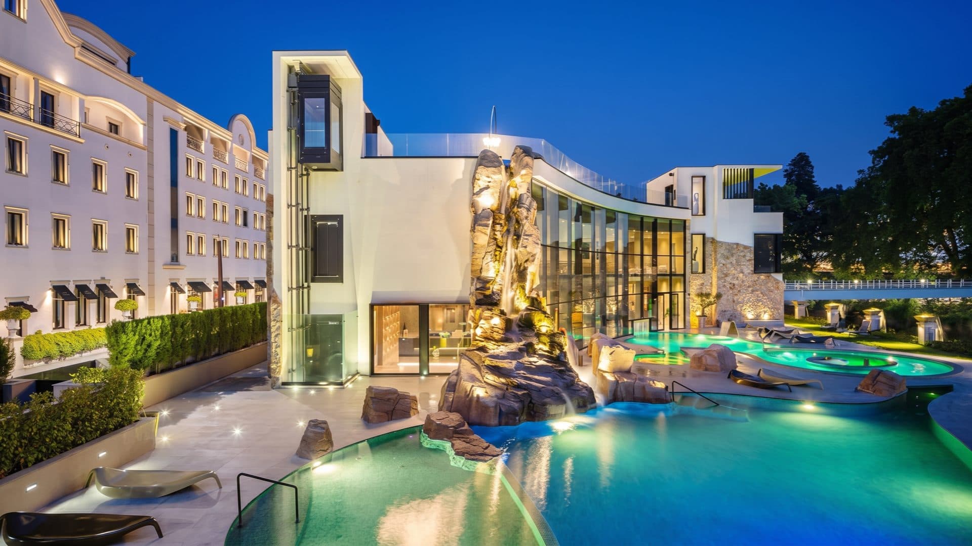 Spa-hotel at dusk, a curved glass wing lit beside a floodlit rock waterfall and turquoise pools, the classical hotel wing alongside.