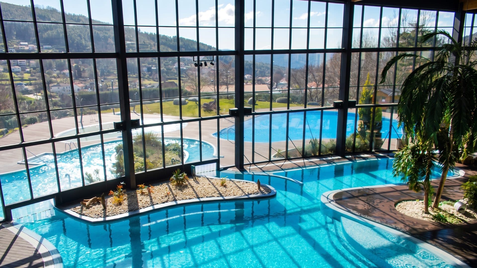 Glass-walled indoor pools with planted gravel islands, tall windows looking out over the mountain valley and the outdoor pools.