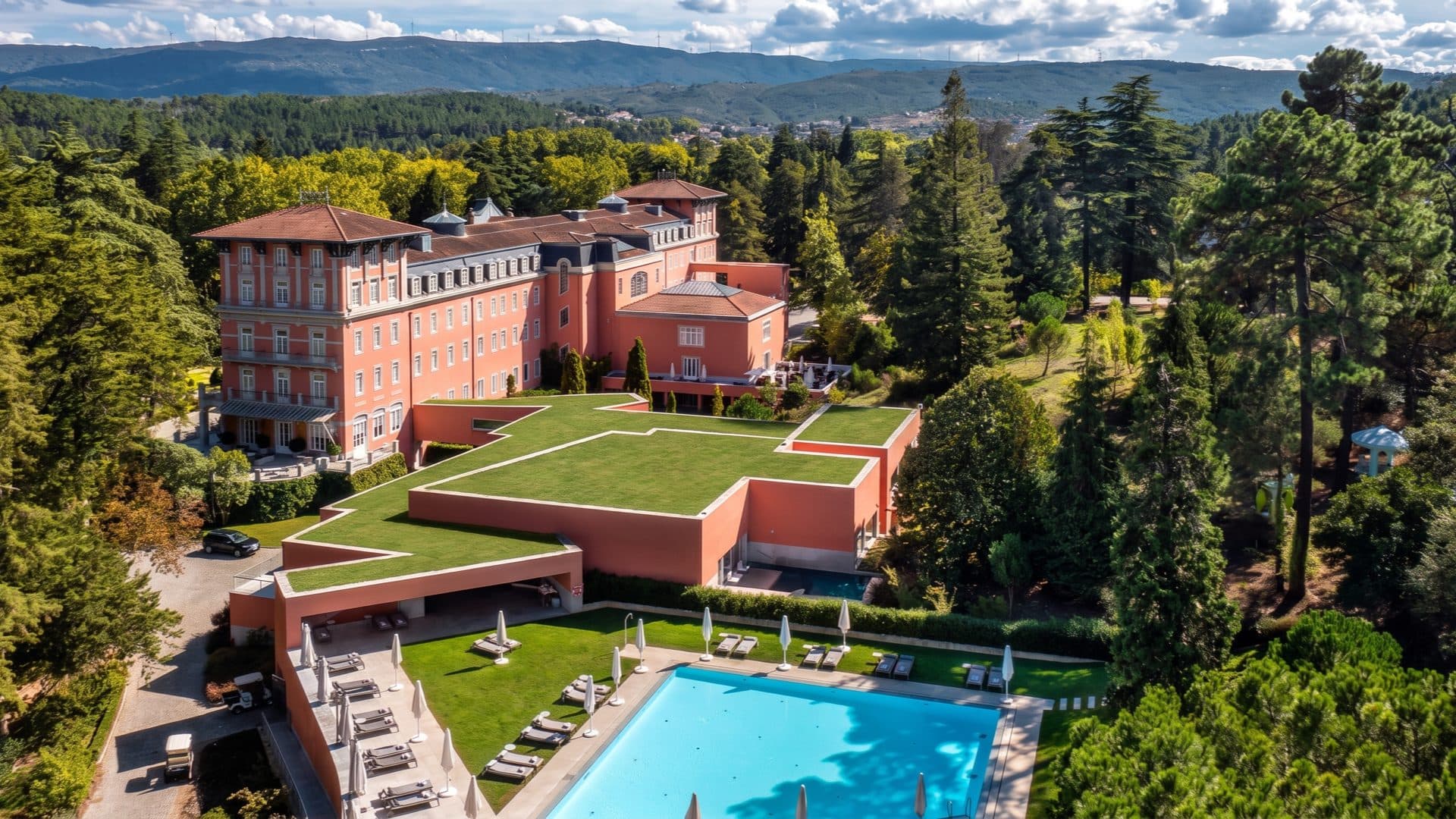 Aerial view of the salmon-pink Vidago Palace hotel among forested parkland, with an outdoor pool and lawns below.