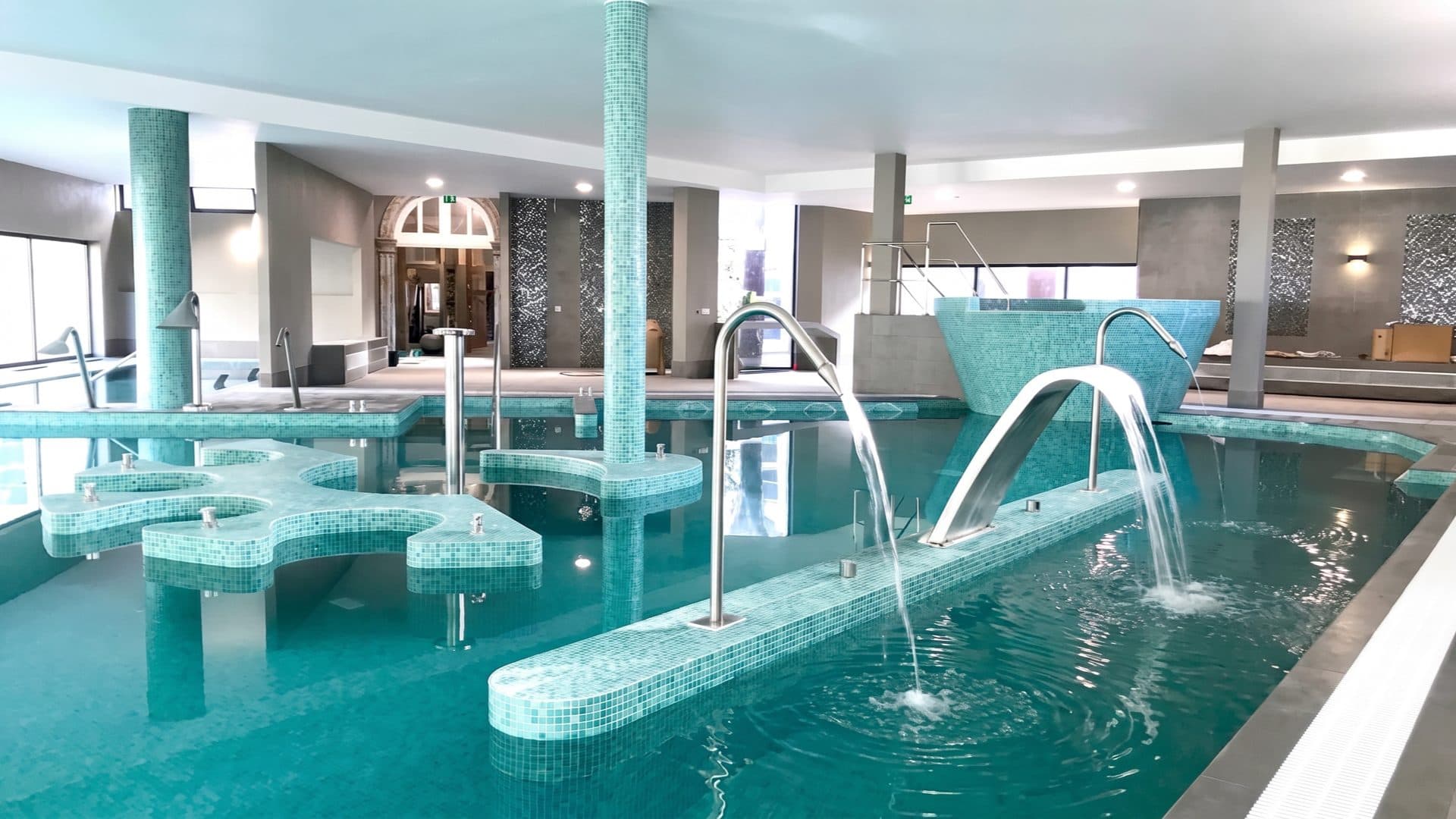 Indoor thermal pool tiled in turquoise mosaic, with hydro jets, slim columns and a historic arched doorway behind.