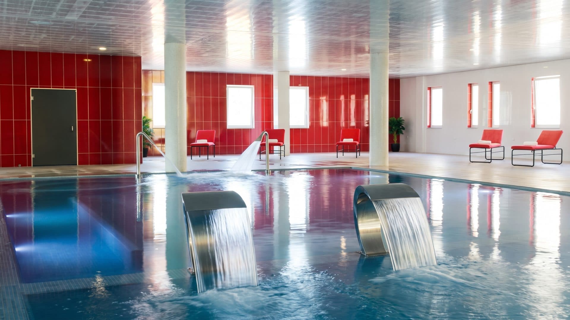 Indoor thermal pool with two steel water blades against red-tiled walls, white columns and red loungers around the deck.