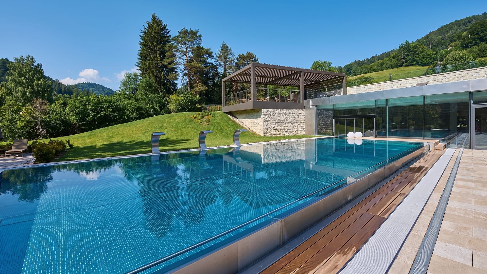 The outdoor thermal pool at Terme di Comano, its overflow edge above the meadow with wooded slopes behind.