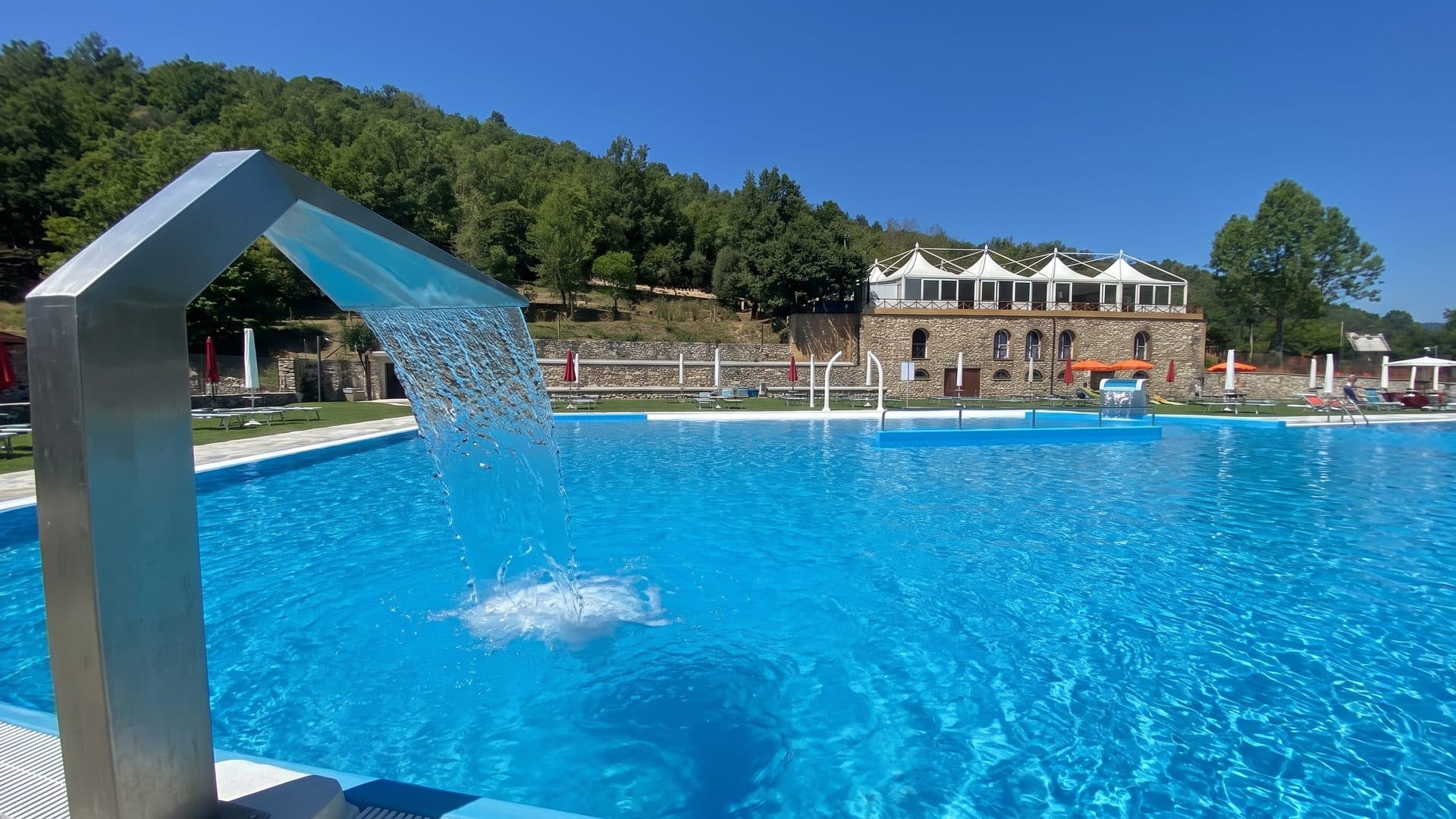 The outdoor thermal pool of Terme di Cretone with its steel waterfall and the arcaded pavilion behind