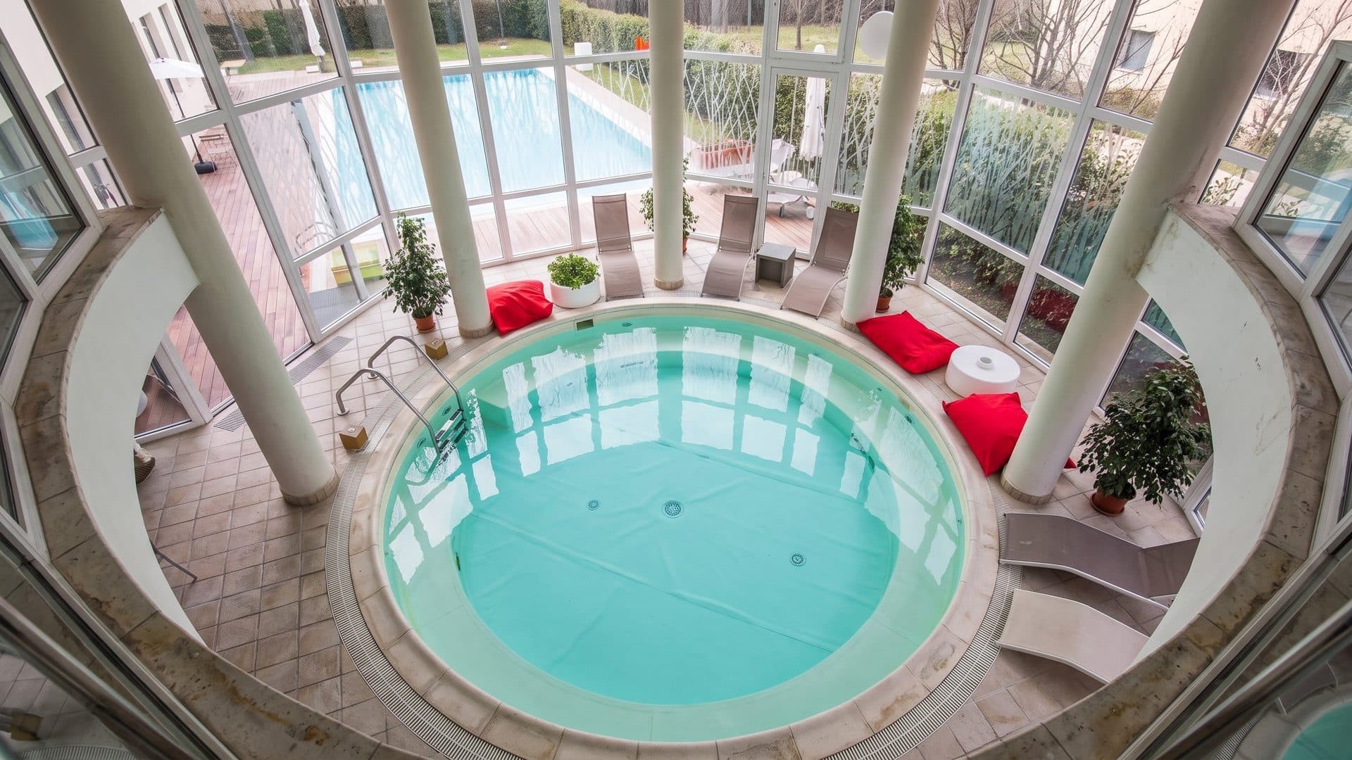 The round indoor thermal pool of the Terme di Gambassi among white columns, with red cushions along the glass wall